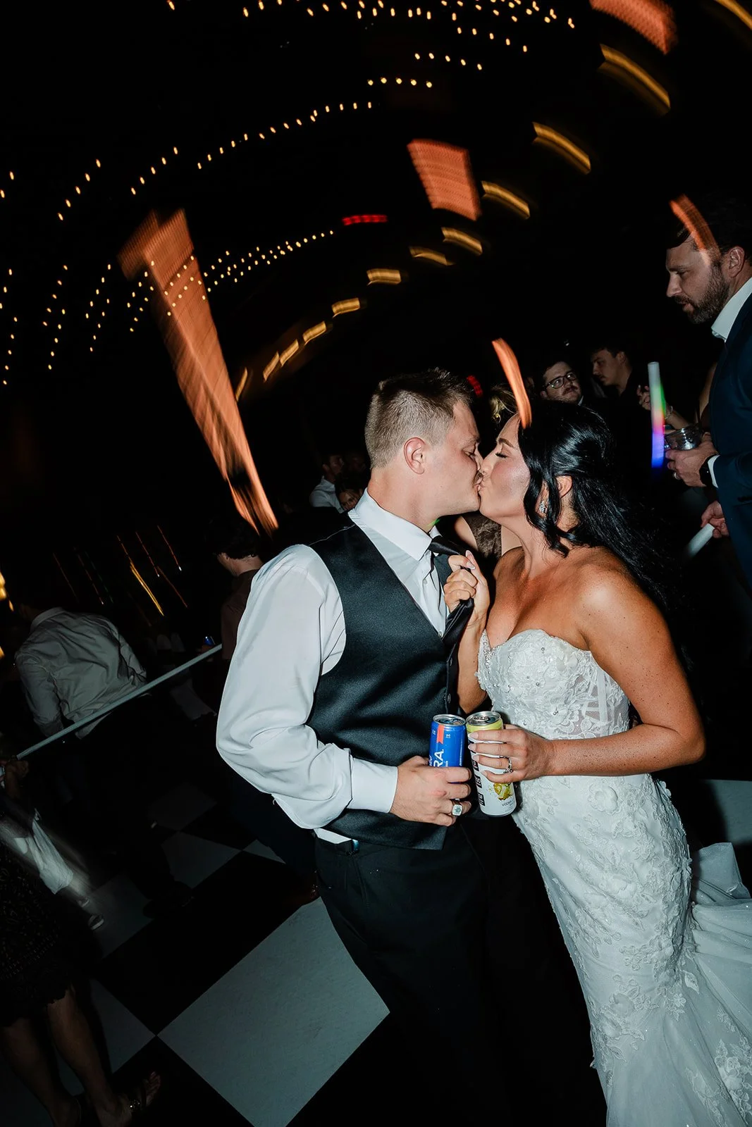 A bride and groom sharing a kiss at their wedding reception, holding cans of drinks, with guests in the background and string lights overhead.