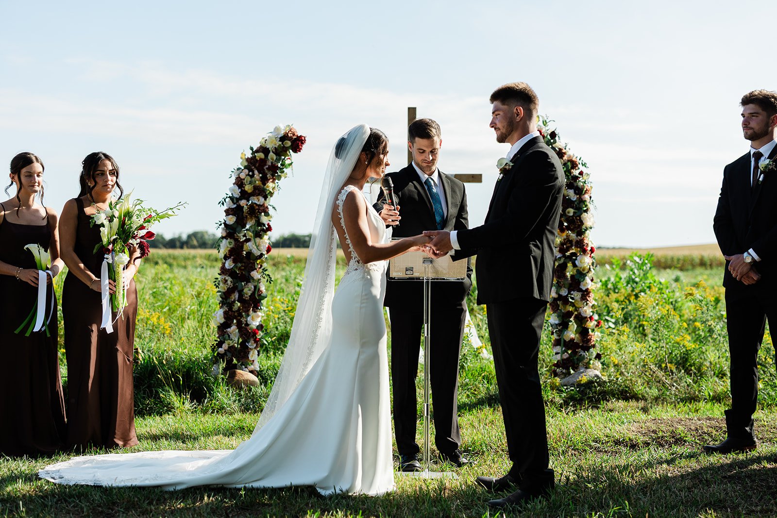 A couple getting married outdoors with wedding officiant and bridesmaids in the background, with a floral arch and green fields in the distance.
