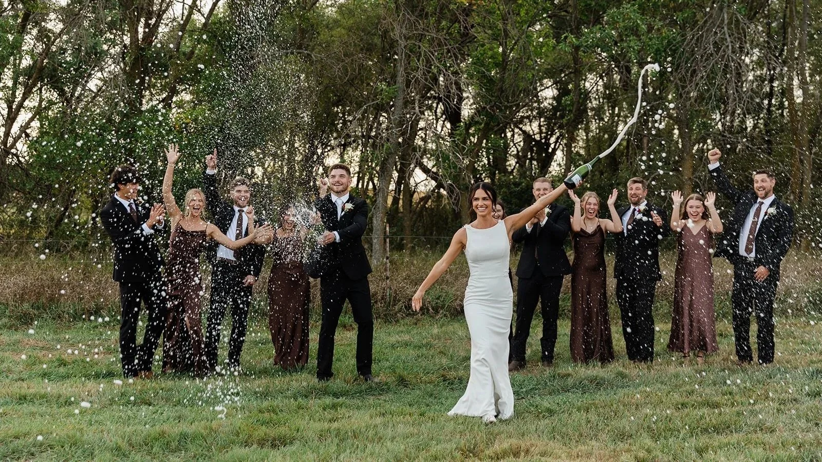 Bride in white dress holding champagne bottle, opening it to celebrate with wedding guests in formal attire outdoors.