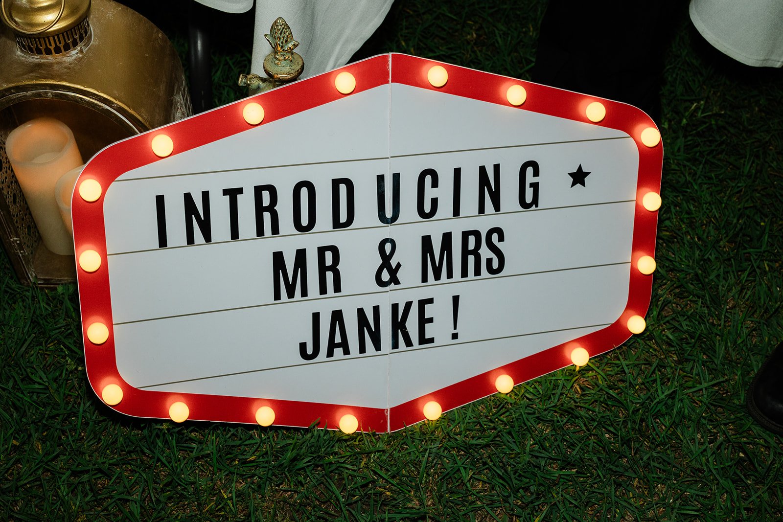 Illuminated sign at a wedding reception introducing Mr. and Mrs. Janke with a border of light bulbs, placed on grass surrounded by candles and decorative items.