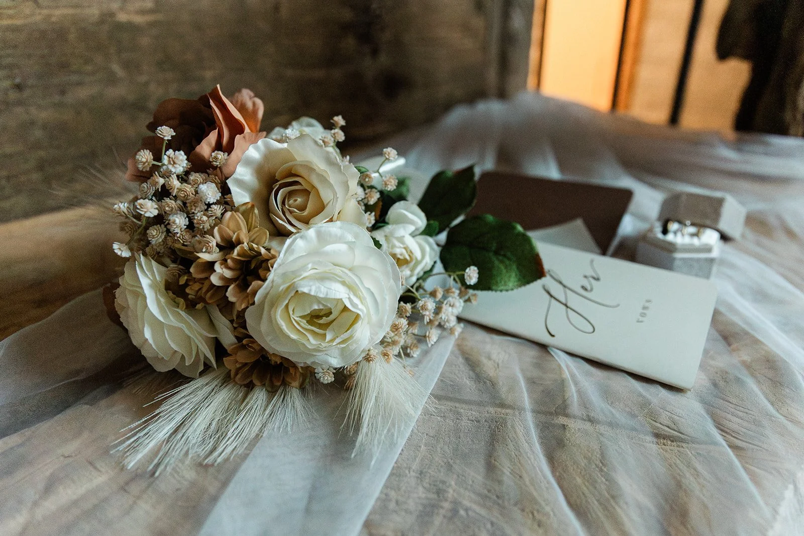 A wedding bouquet with white roses, small white flowers, and brown accents, placed on a sheer fabric, with a letter, a jewelry box, and a brick wall background.