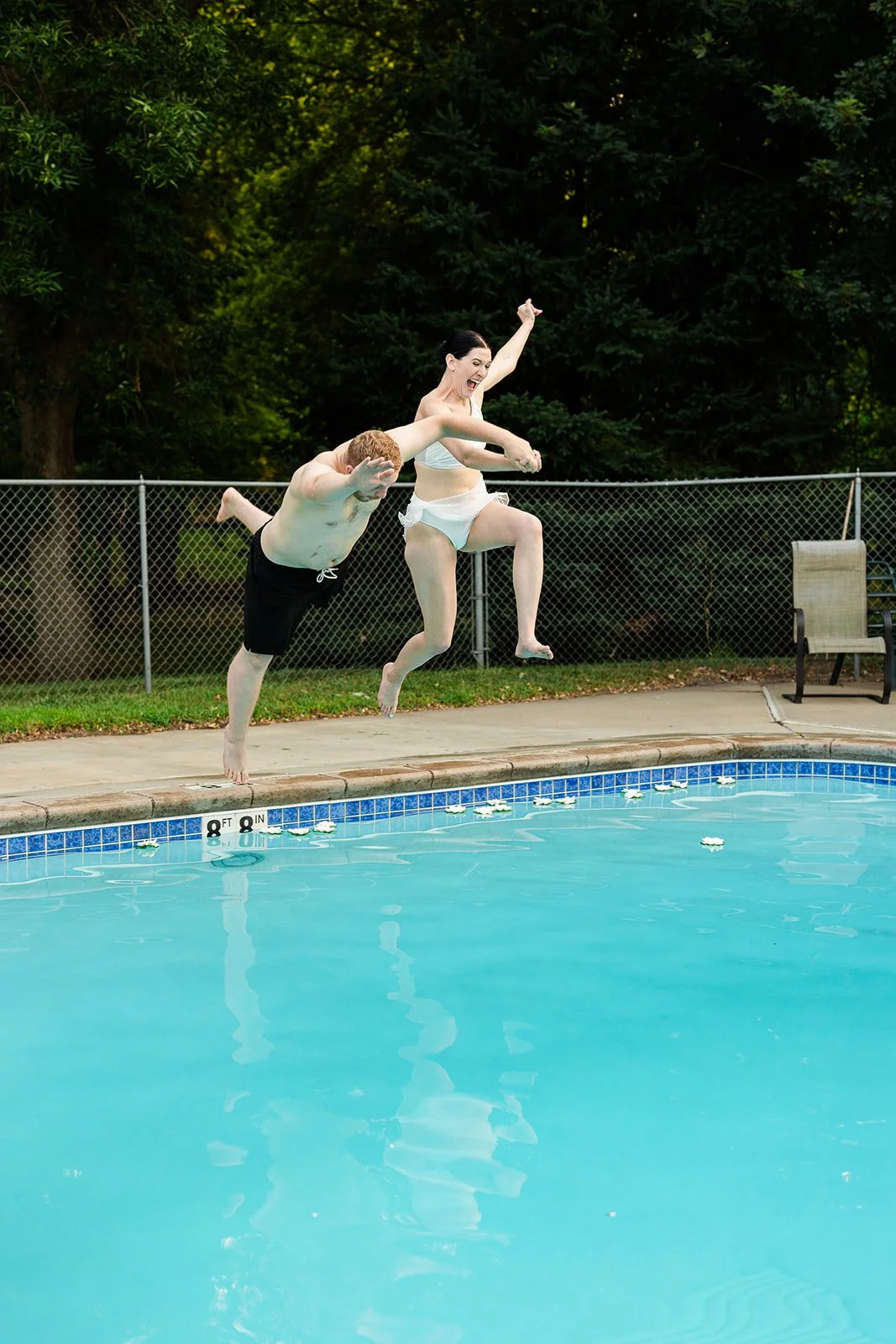 A man and woman jumping into a swimming pool from the poolside, with the woman wearing a white swimsuit and the man shirtless in swim shorts, surrounded by a backyard with trees and a fence.