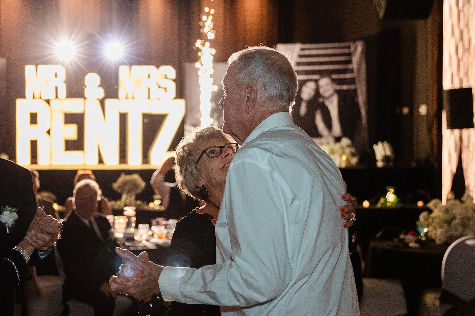 An elderly couple dancing closely at a wedding reception, with a large illuminated sign in the background reading "Mr & Mrs Rentyz" and wedding guests seated at tables.