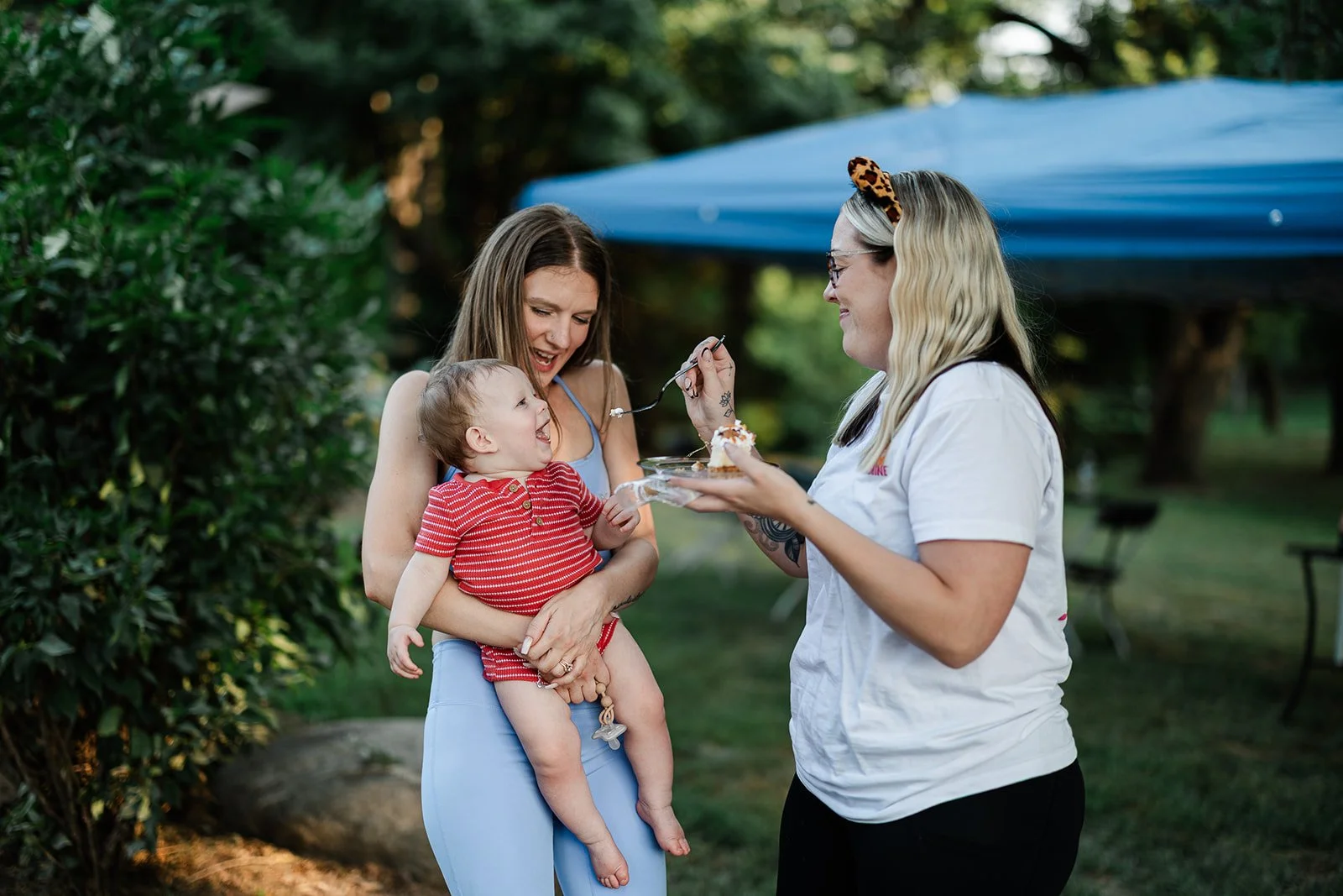 A woman holding a smiling toddler in her arms while another woman feeds them cake outdoors during daytime.