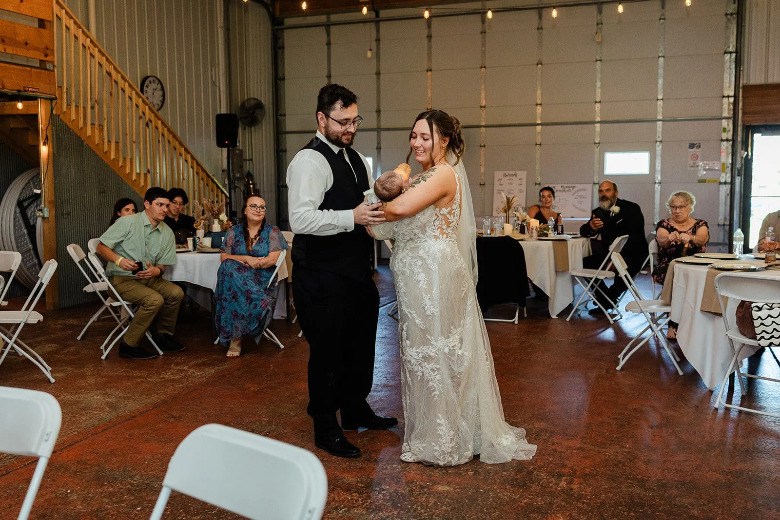 A wedding reception with a bride and groom holding a newborn baby in an industrial-style venue. Guests are seated at round tables, watching the couple with smiles. The bride wears a lace wedding gown, and the groom wears a black vest and white shirt.