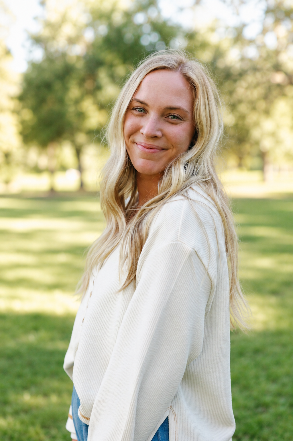 A young woman with long blonde hair smiling outdoors in a park with green trees and sunlight.