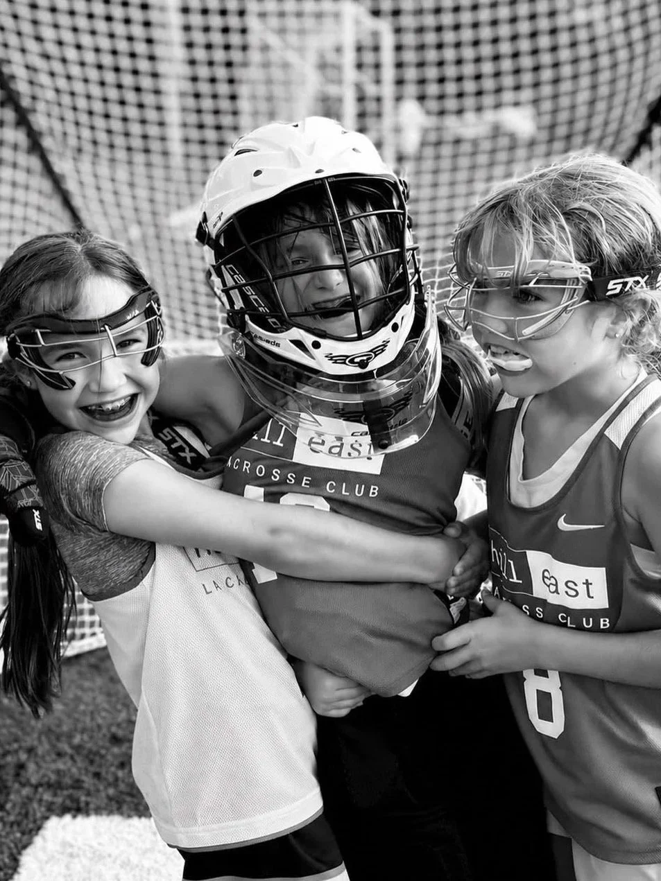 Three young girls in sports gear, wearing helmets and goggles, smiling and embracing each other on a lacrosse field.
