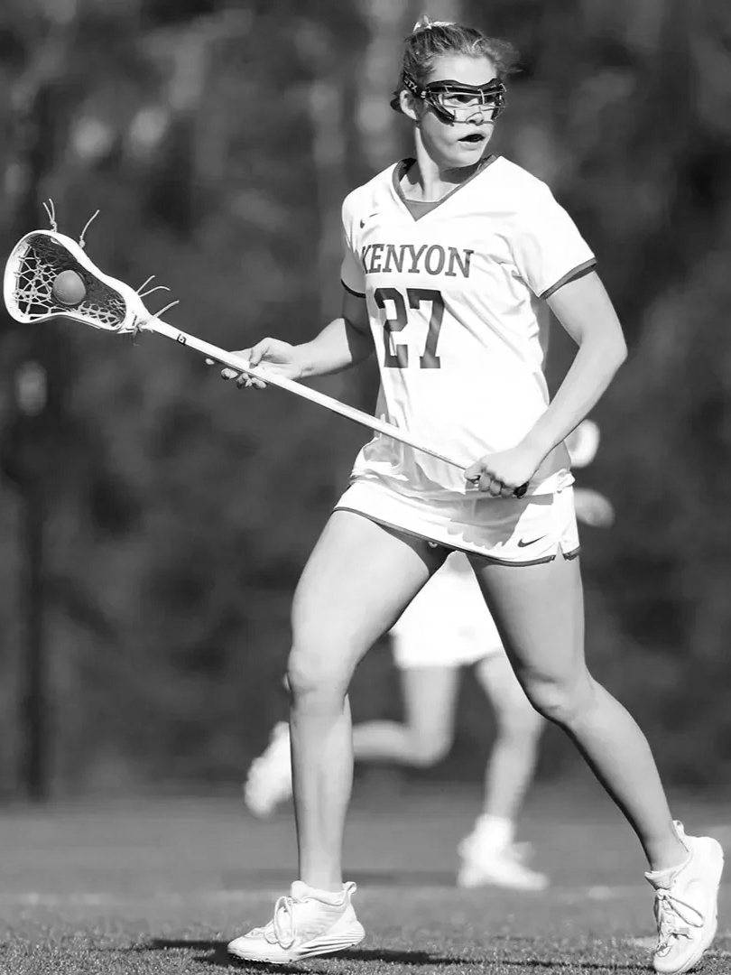 A female lacrosse player in uniform with the number 27, holding a lacrosse stick with a ball, on the field during a game, wearing protective goggles.