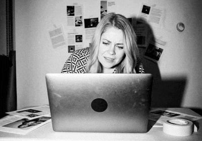 A woman sitting at a desk in front of a laptop, appearing frustrated or upset, with papers and tape on the desk and photos or notes on the wall behind her.