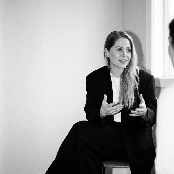 A woman with long hair, wearing a black blazer and pants, sitting on a chair by a window, engaged in a conversation, gesturing with her hands.