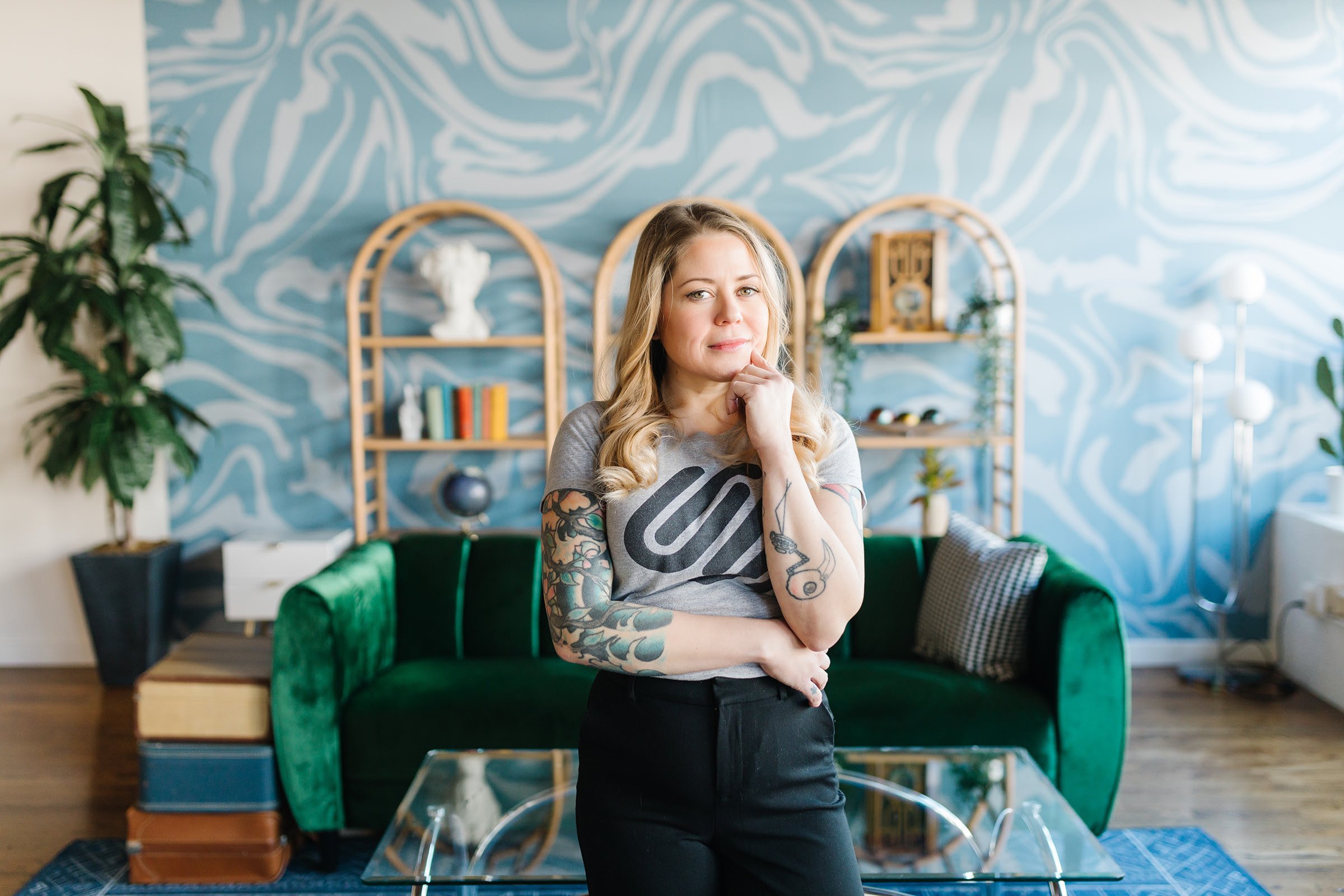 A woman with tattoos standing in a modern, stylish living room with a blue abstract wallpaper, green velvet couch, and decorative shelves with books and ornaments.