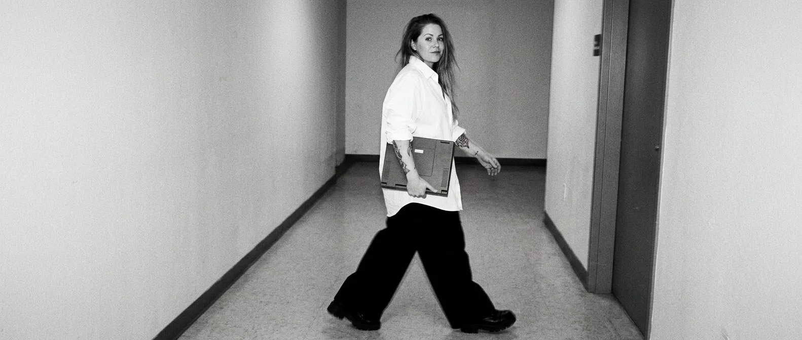 A woman with long hair, wearing a white shirt and black pants, walking down a hallway while holding a laptop, looking at the camera.