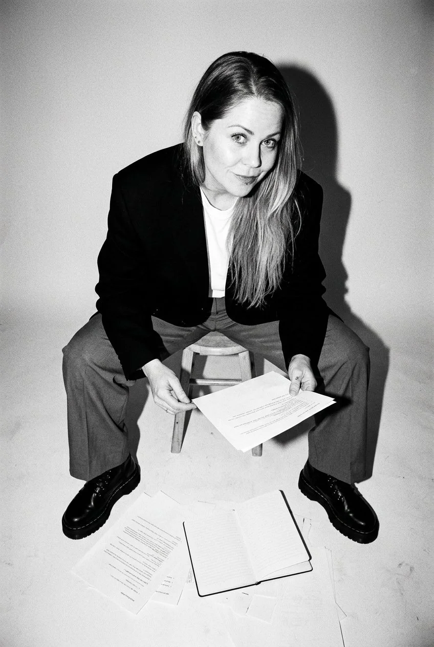 A woman with long hair, dressed in a black blazer and light-colored pants, is sitting on a small wooden stool. She is holding a piece of paper and has documents and an open notebook on the floor in front of her. The photo is in black and white.