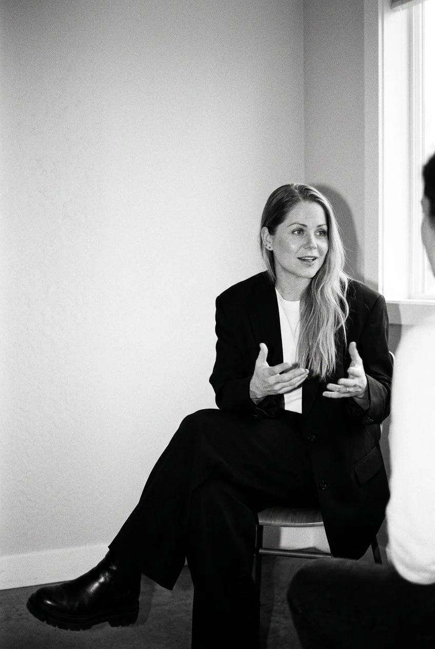 A woman with long hair talking to a person whose back is to the camera in a room with a window, wearing a blazer and sitting on a chair.