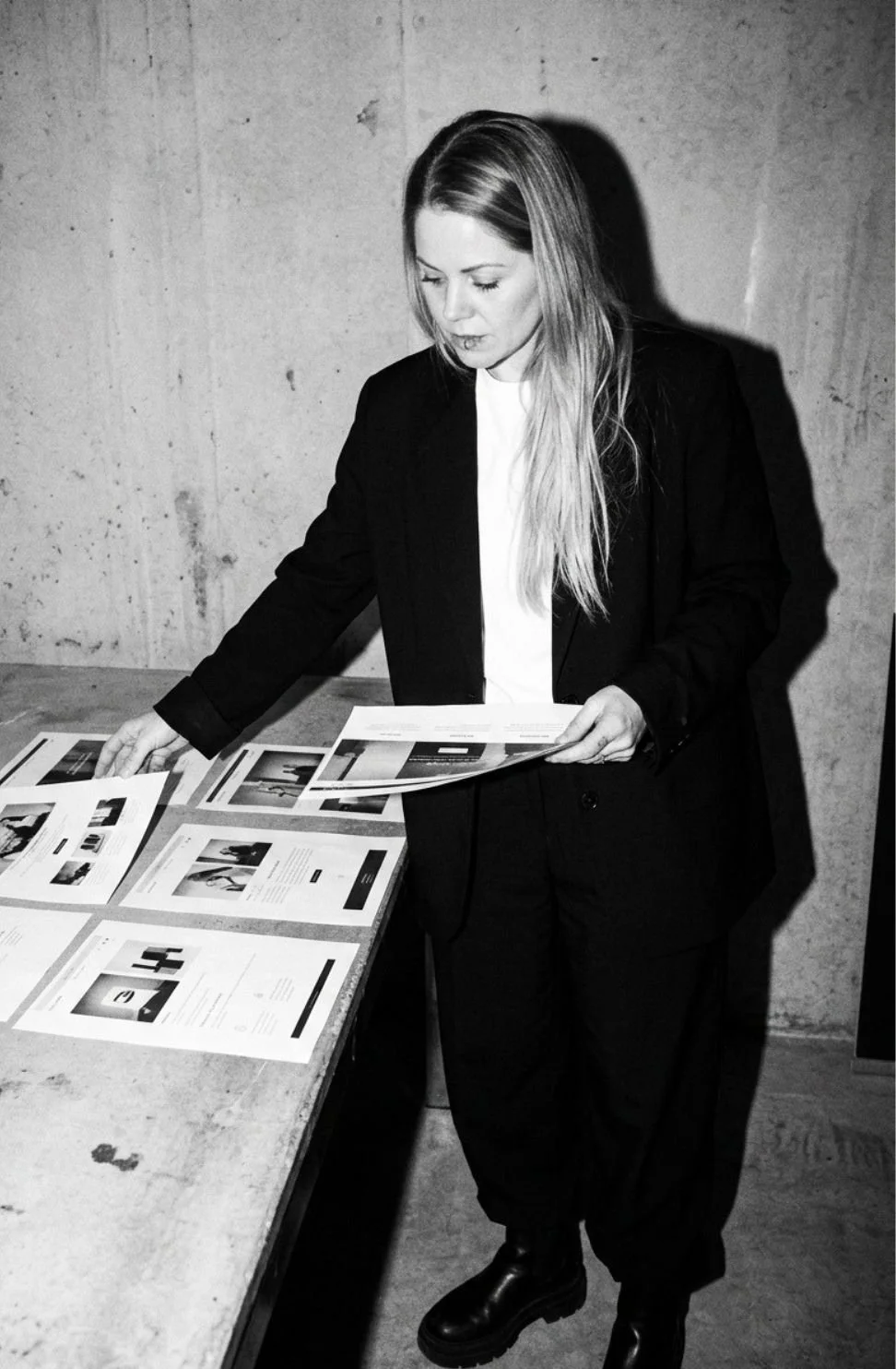 A woman with long hair dressed in a black suit and white shirt looking at printed documents or brochures on a table in an industrial-style room.