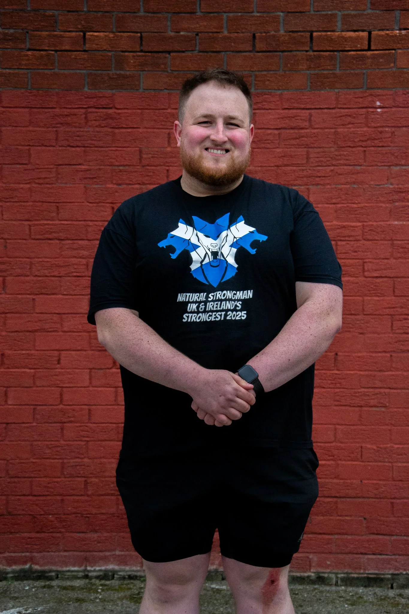 Smiling man in a black strongman T-shirt standing with his hands clasped against a red brick wall.