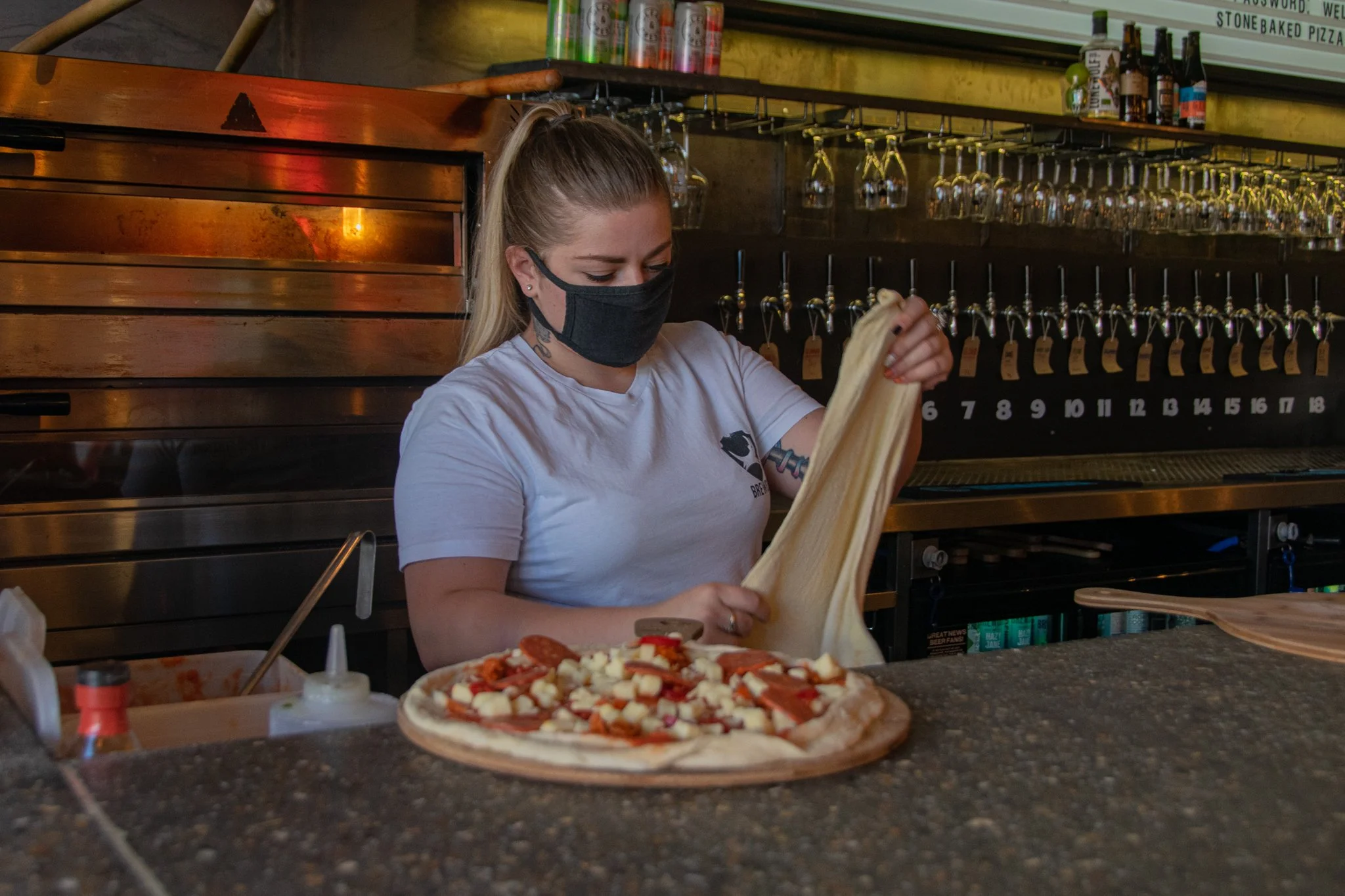 Person stretching pizza dough behind a counter, with a topped pizza in the foreground.