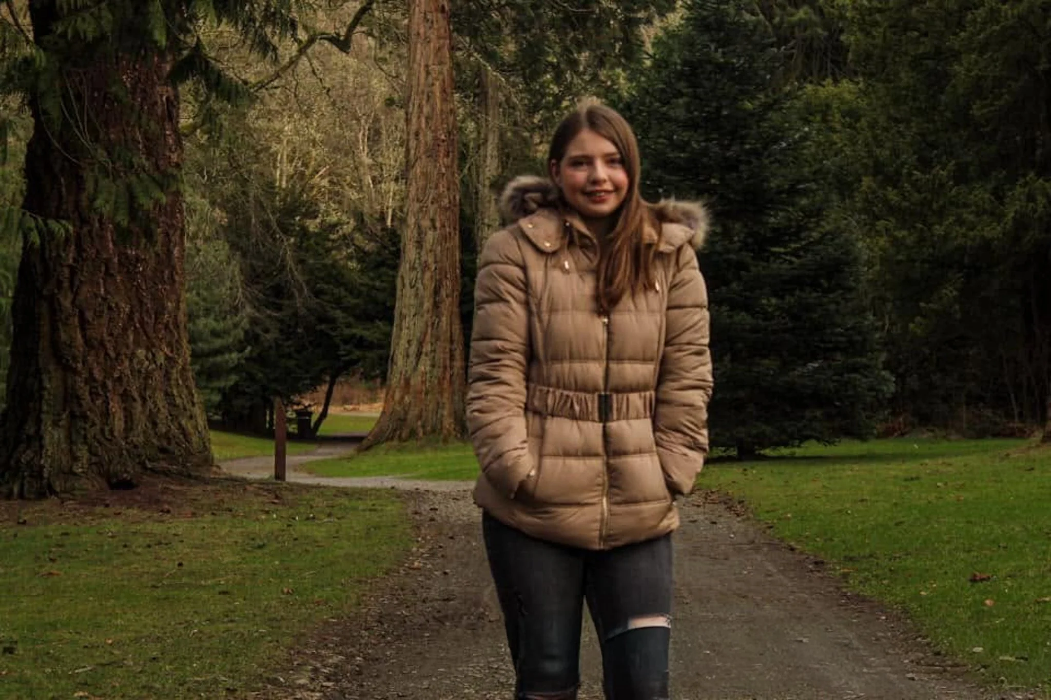 Outdoor self-portrait of Jade wearing a tan coat on a tree-lined path.
