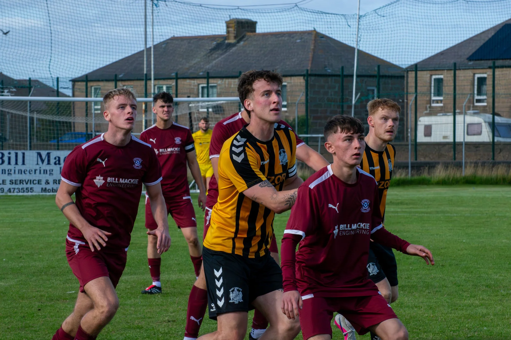 Footballers in maroon and yellow-and-black kits looking upwards as they anticipate the ball.