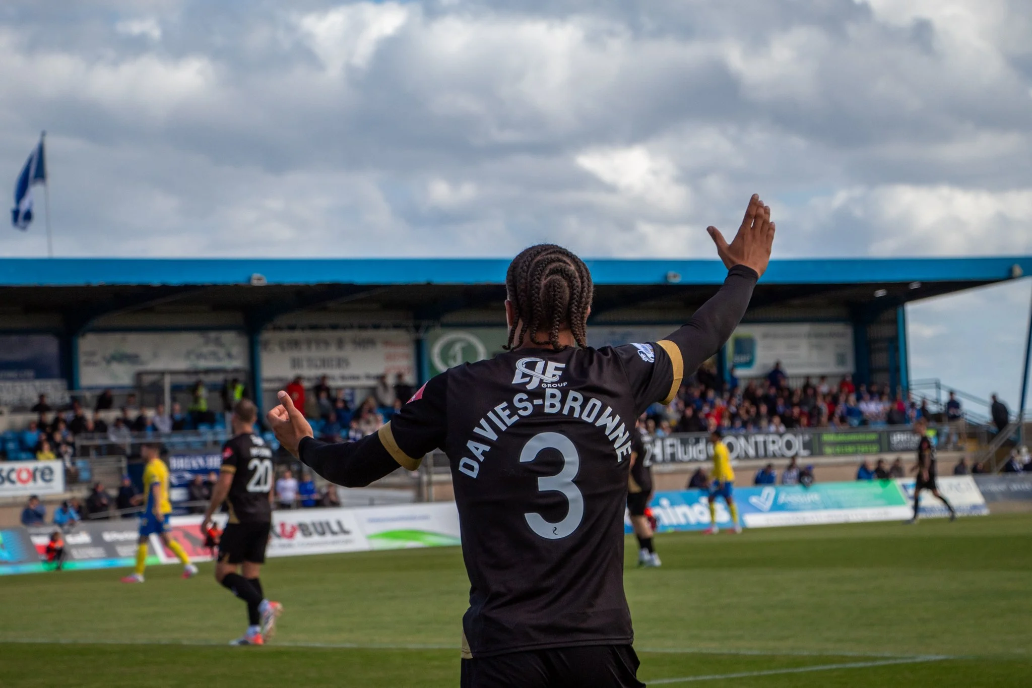 Footballer wearing a black number 3 shirt raising his arms, with spectators in the stand behind him.
