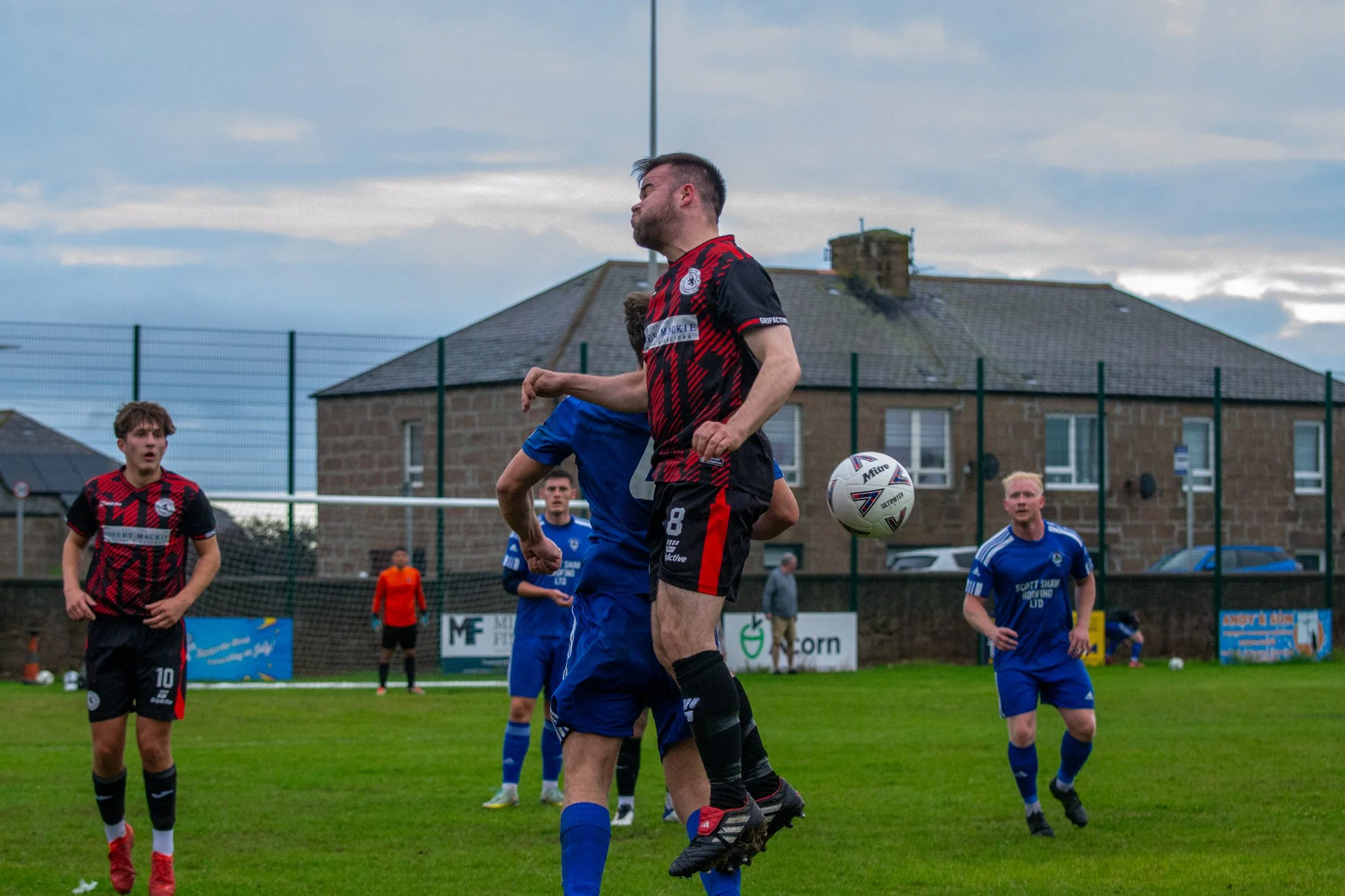 Footballers in red-and-black and blue kits competing for the ball in the air.