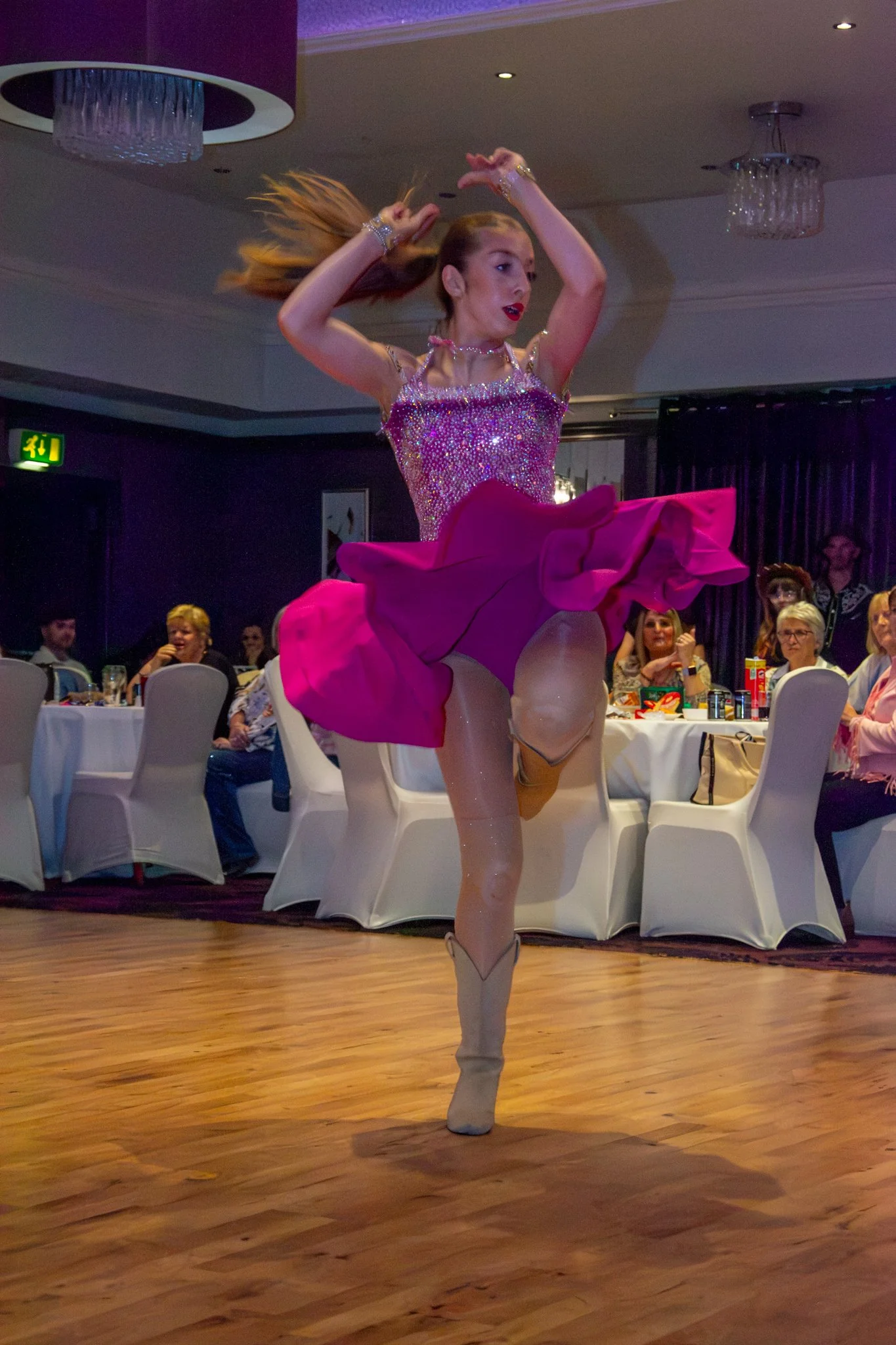 Dancer in a bright pink dress performing with her arms raised, with guests seated behind her.
