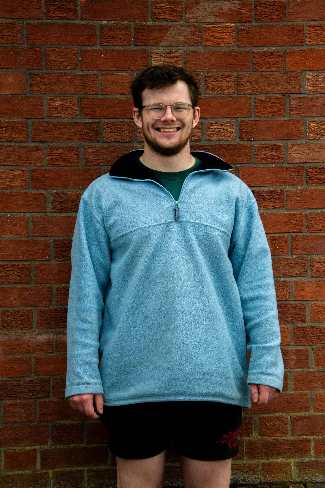 Smiling man wearing glasses and a pale blue fleece against a red brick wall.