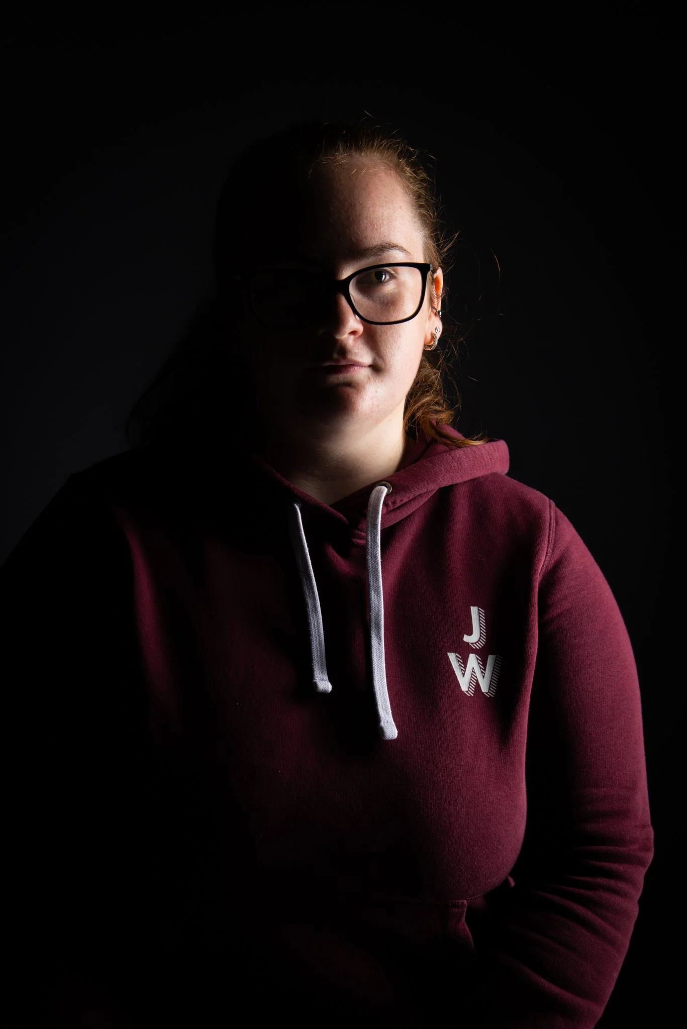 Portrait of a woman in glasses and a burgundy hoodie, with one side of her face lit against a dark background.