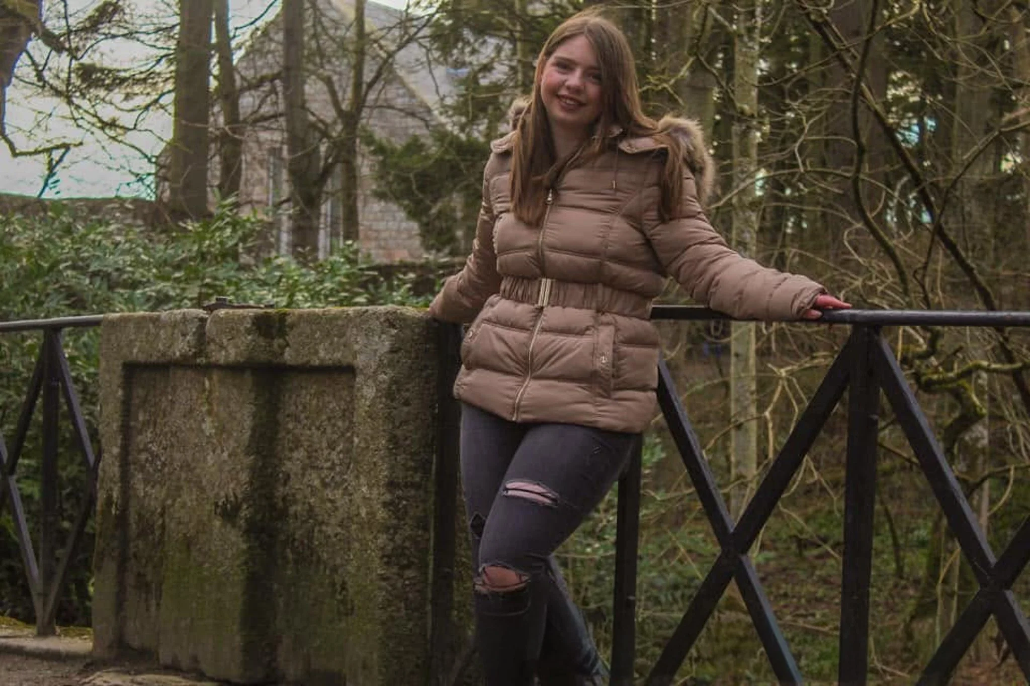 Self-portrait of Jade smiling in a tan coat beside a stone pillar and metal railing, with trees behind her.