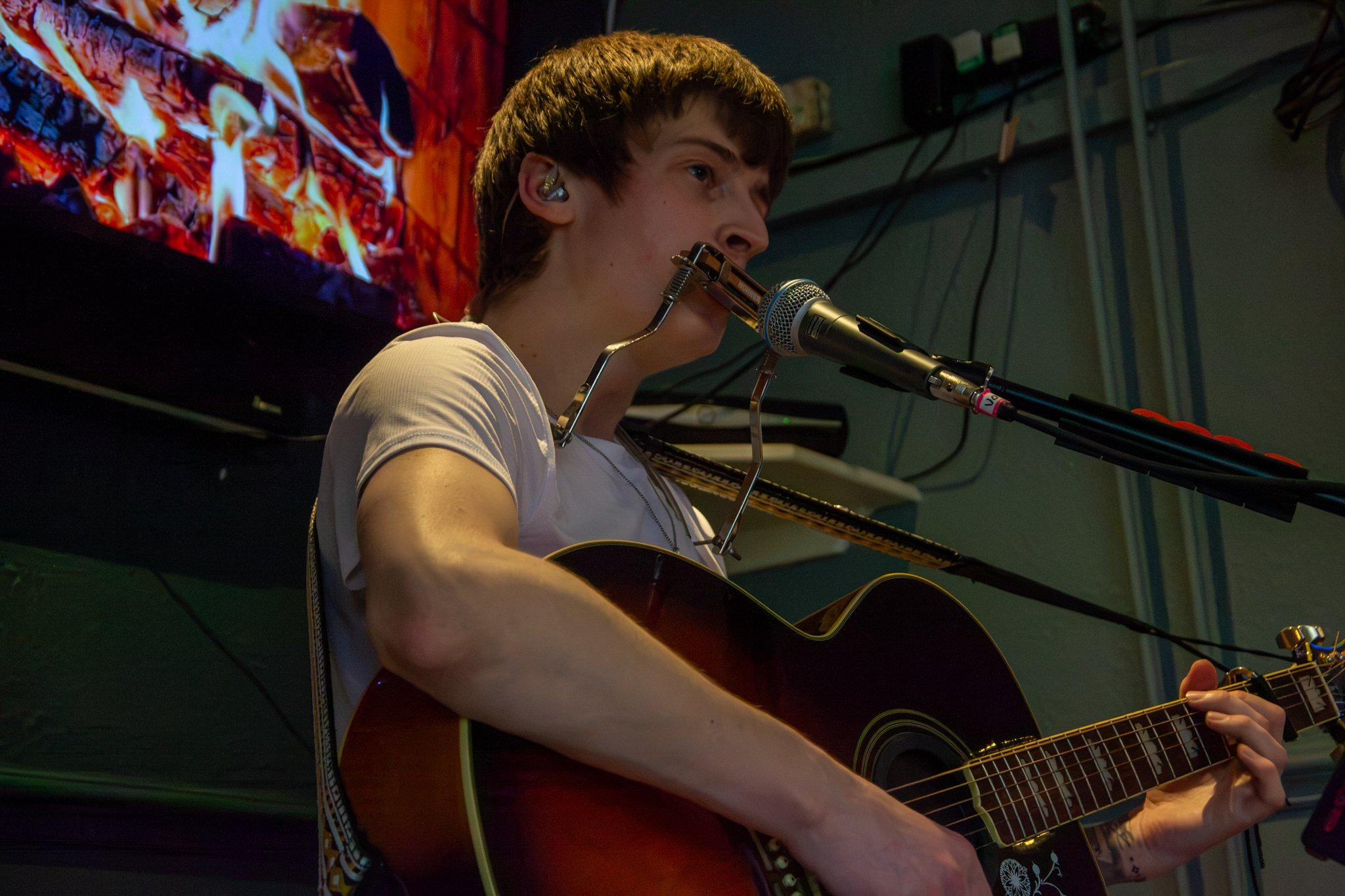 Musician playing an acoustic guitar at a microphone, with a harmonica mounted near his mouth.
