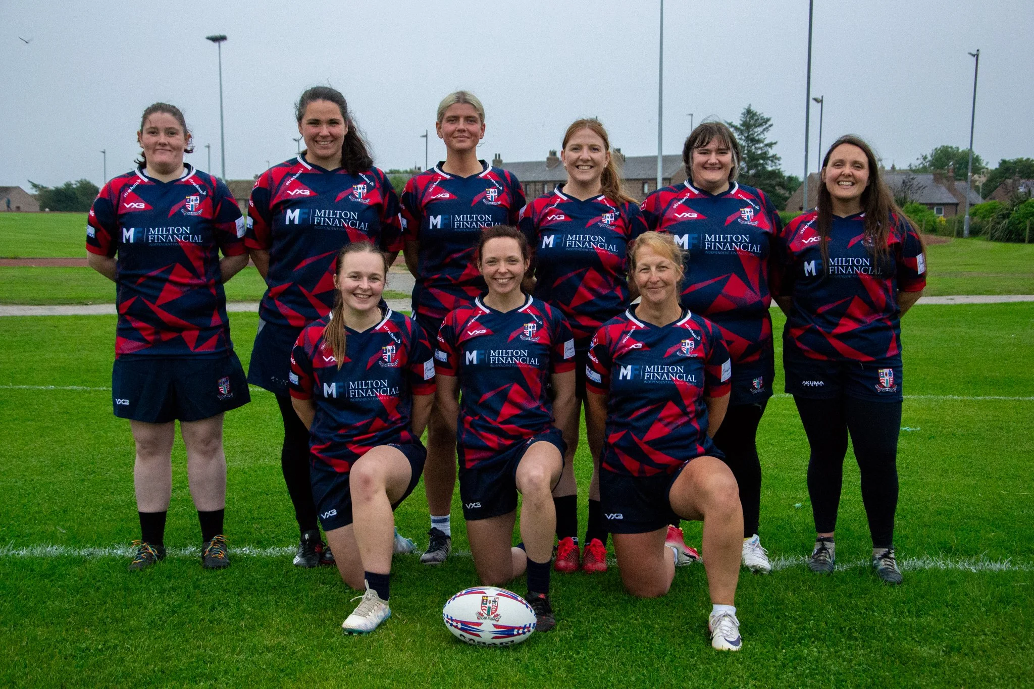Rugby team posing in two rows on a grass pitch, with a rugby ball in front.