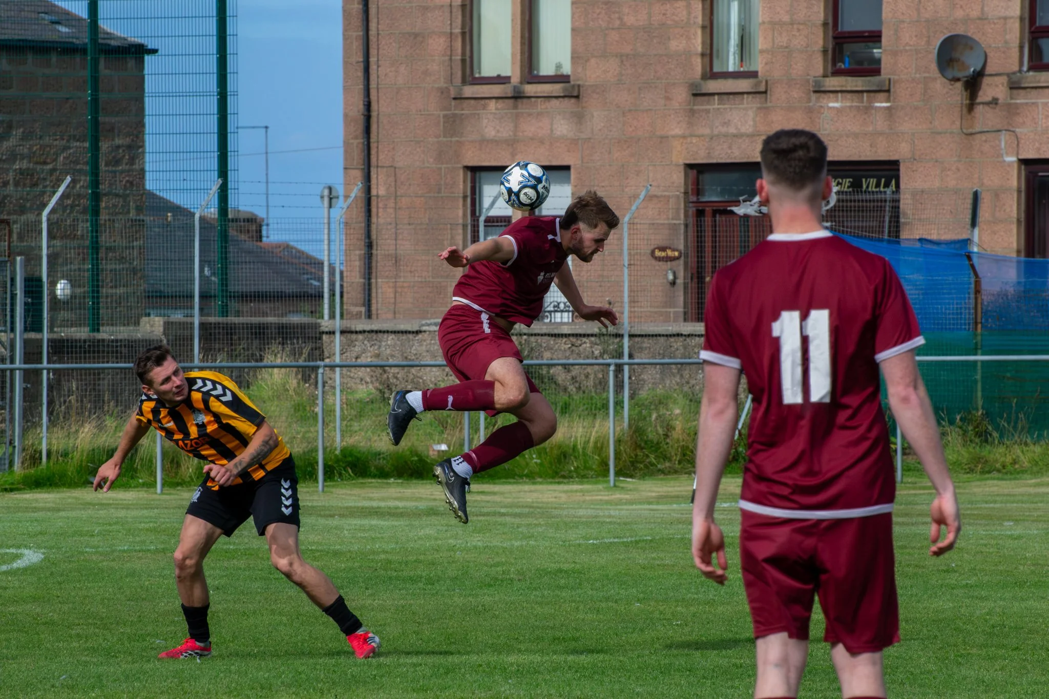 Footballer in maroon jumping to head the ball, with other players nearby on the pitch.