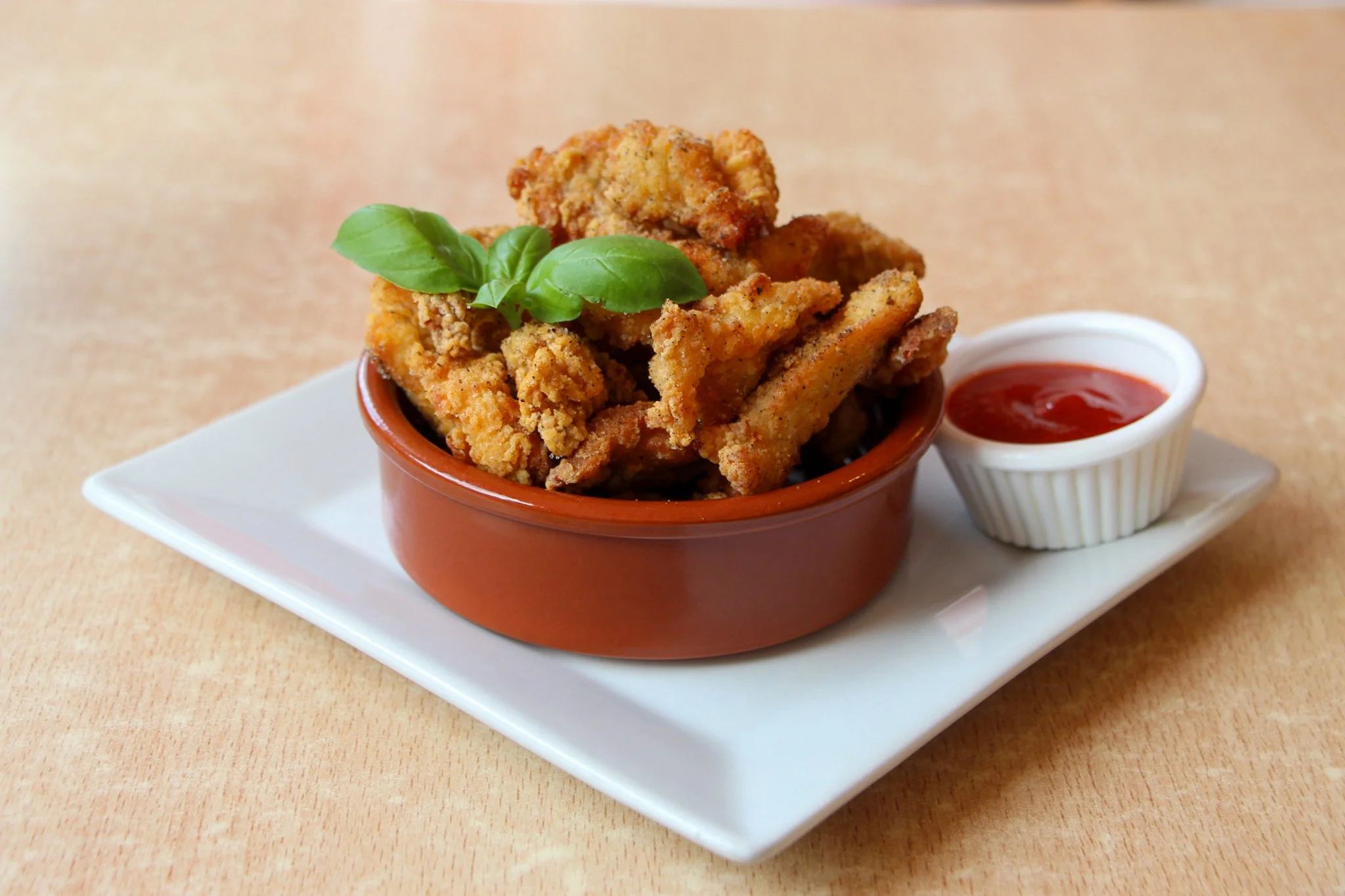 Crispy fried food garnished with basil in a brown bowl, served with a red dipping sauce on a white plate.