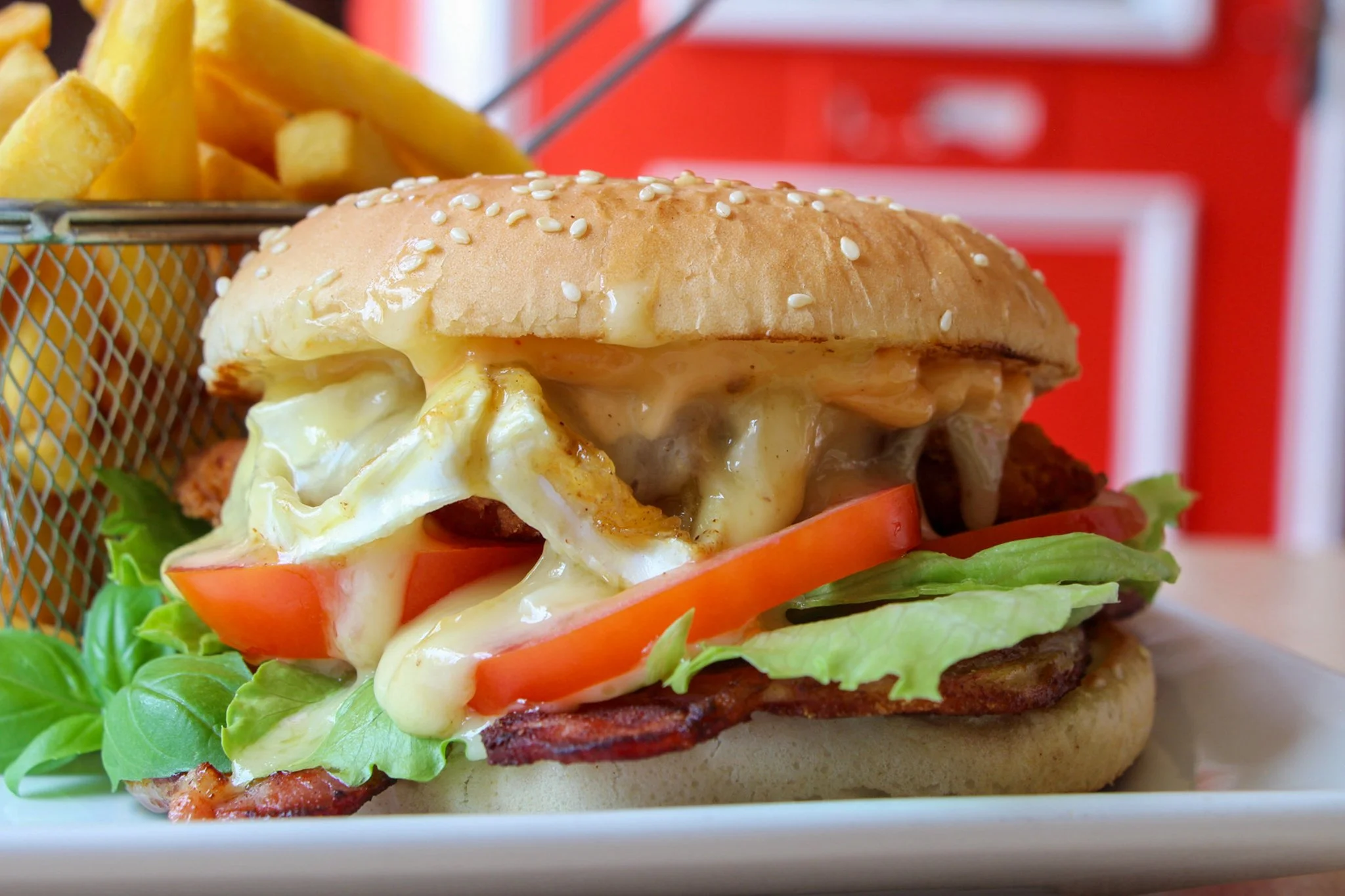 Close-up of a burger with melted cheese, bacon, lettuce and tomato, served beside a basket of chips.