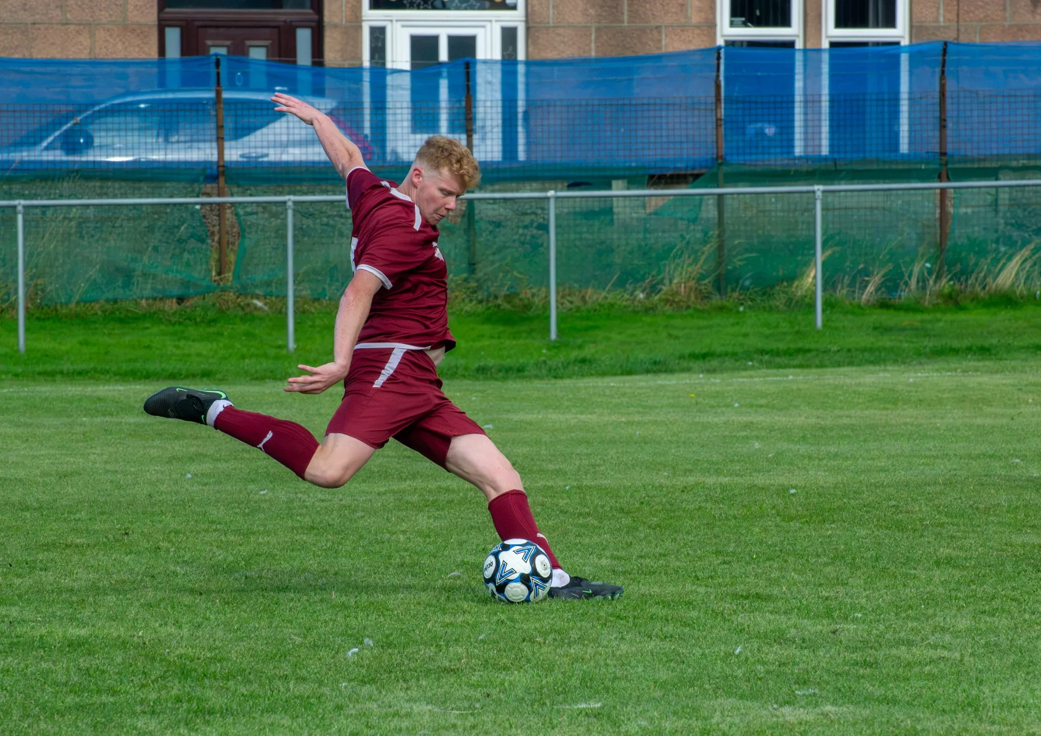 Footballer in maroon swinging his leg to kick the ball on a grass pitch.