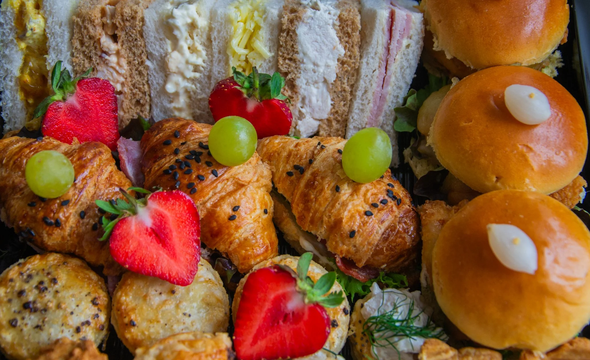 Platter of sandwiches, filled croissants and small rolls, garnished with strawberries and green grapes.