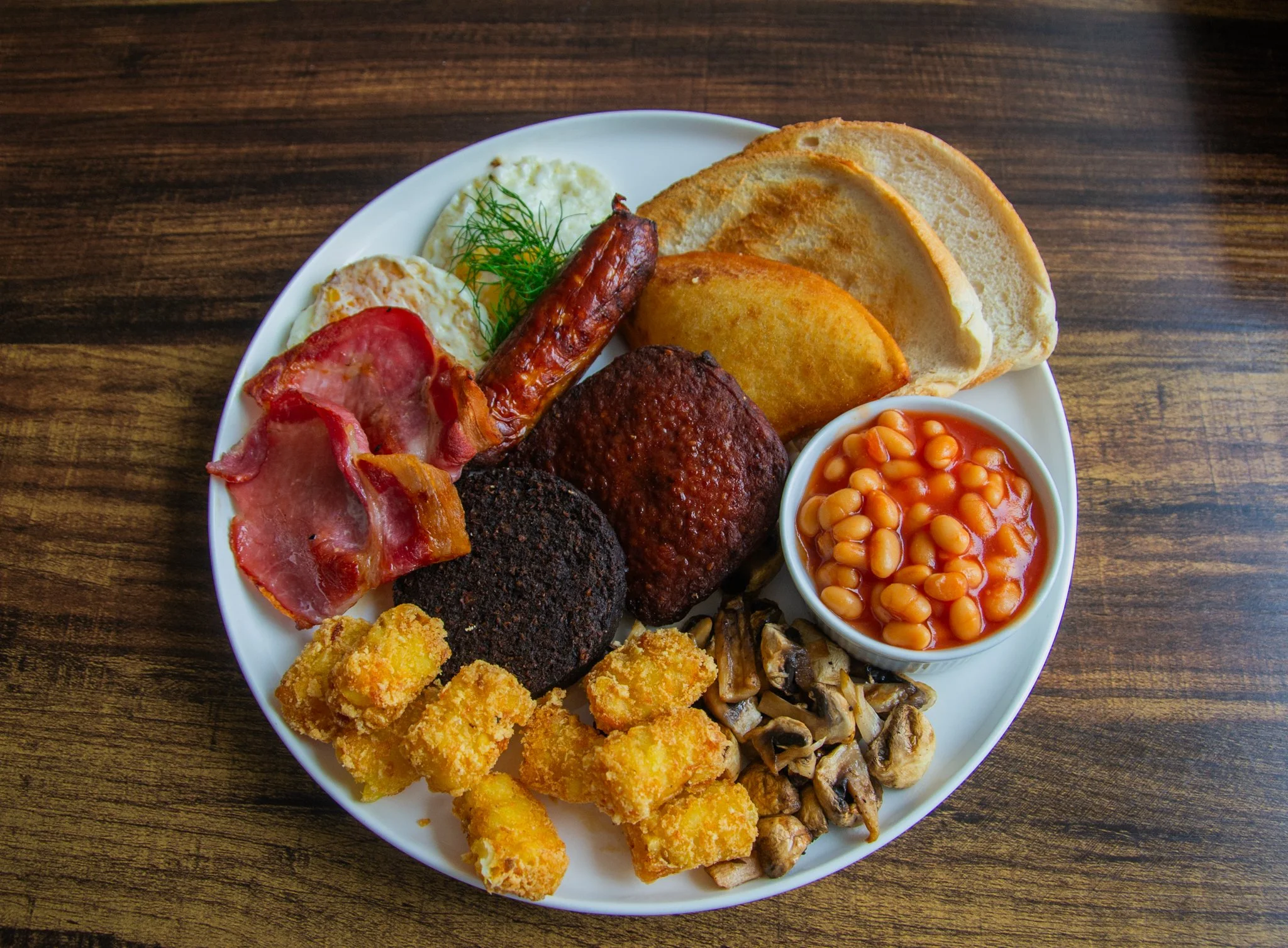 Overhead view of a cooked breakfast with eggs, bacon, sausage, beans, mushrooms, toast and potato bites.