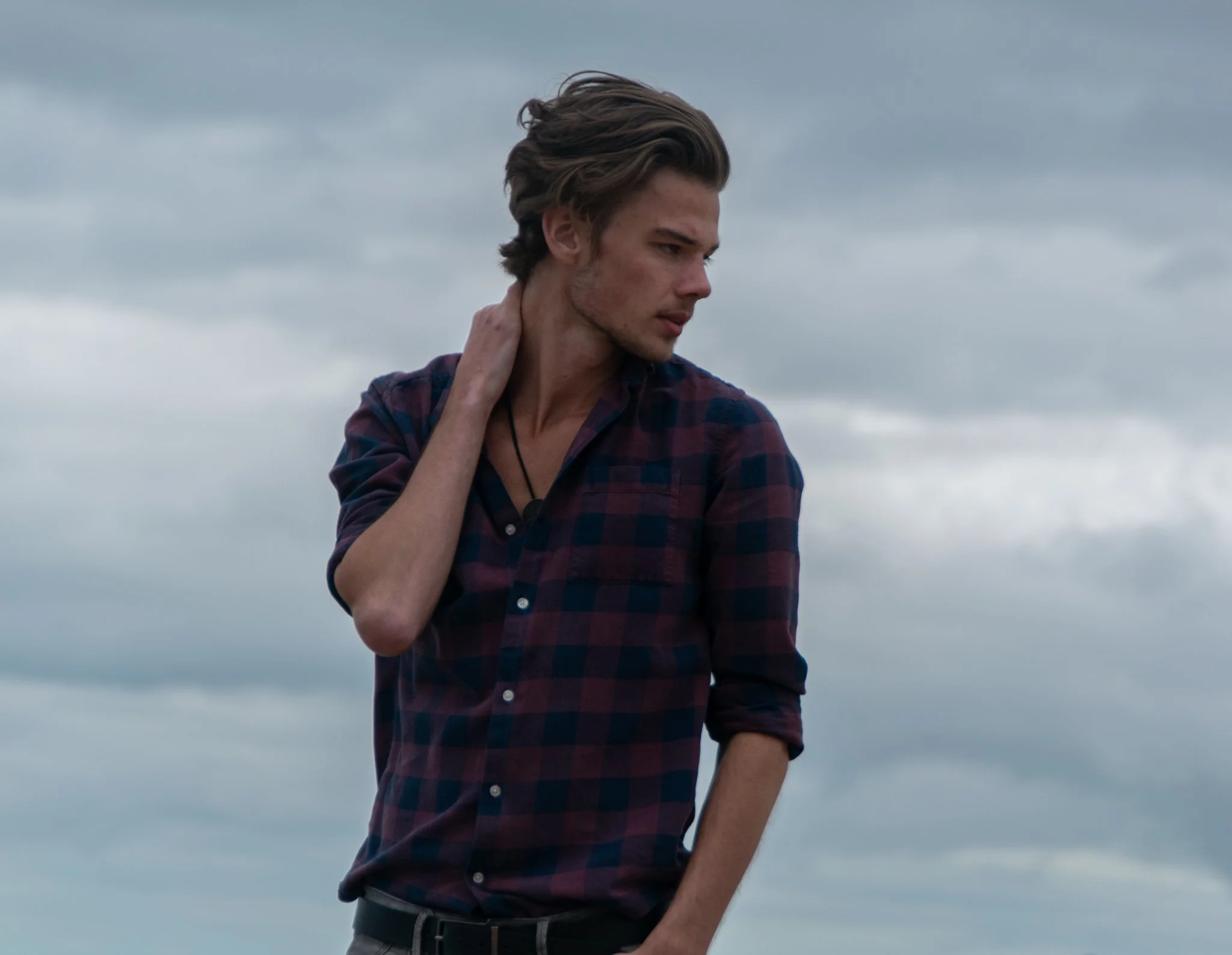 Man in a checked shirt posing outdoors with one hand behind his neck against a cloudy sky.