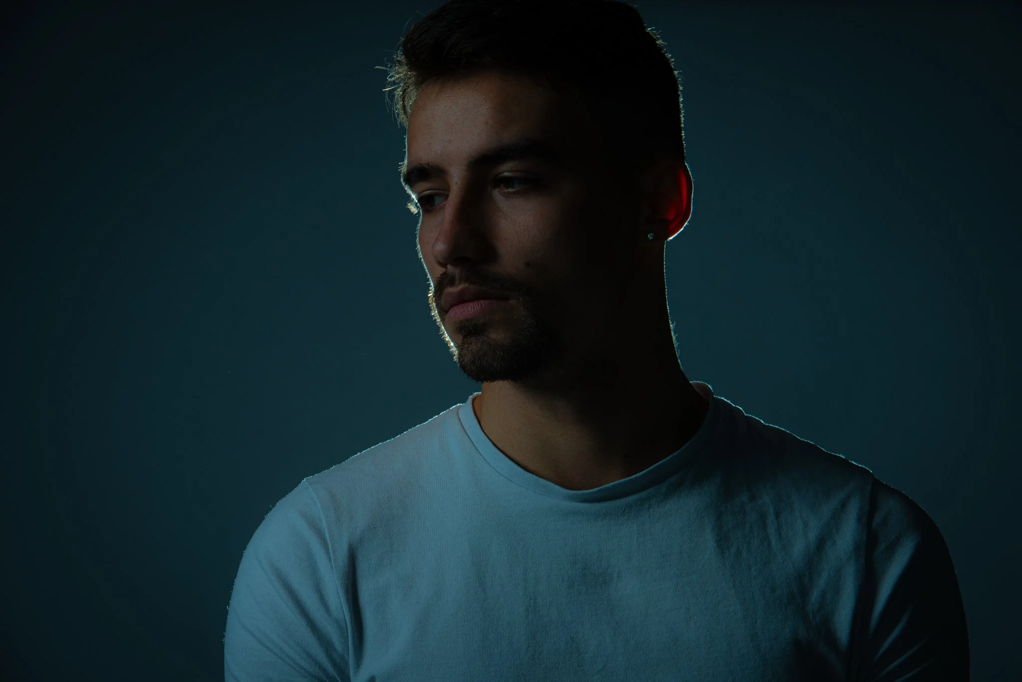 Man in a pale blue T-shirt looking to the side, outlined by light against a dark teal background.