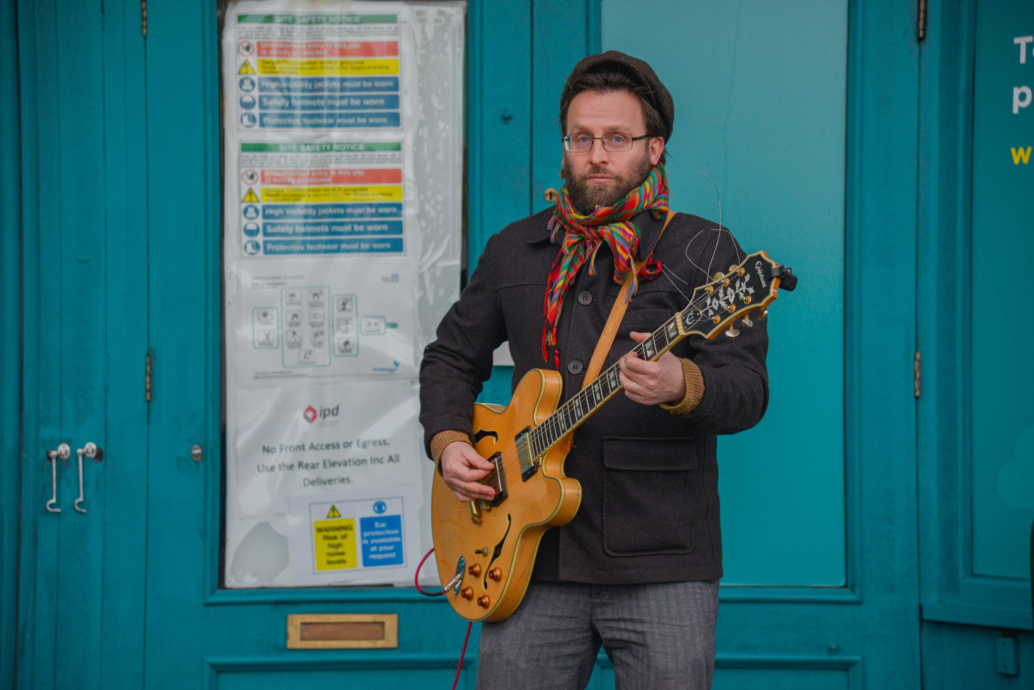 Man wearing glasses, a hat and a colourful scarf, playing a guitar in front of turquoise doors.