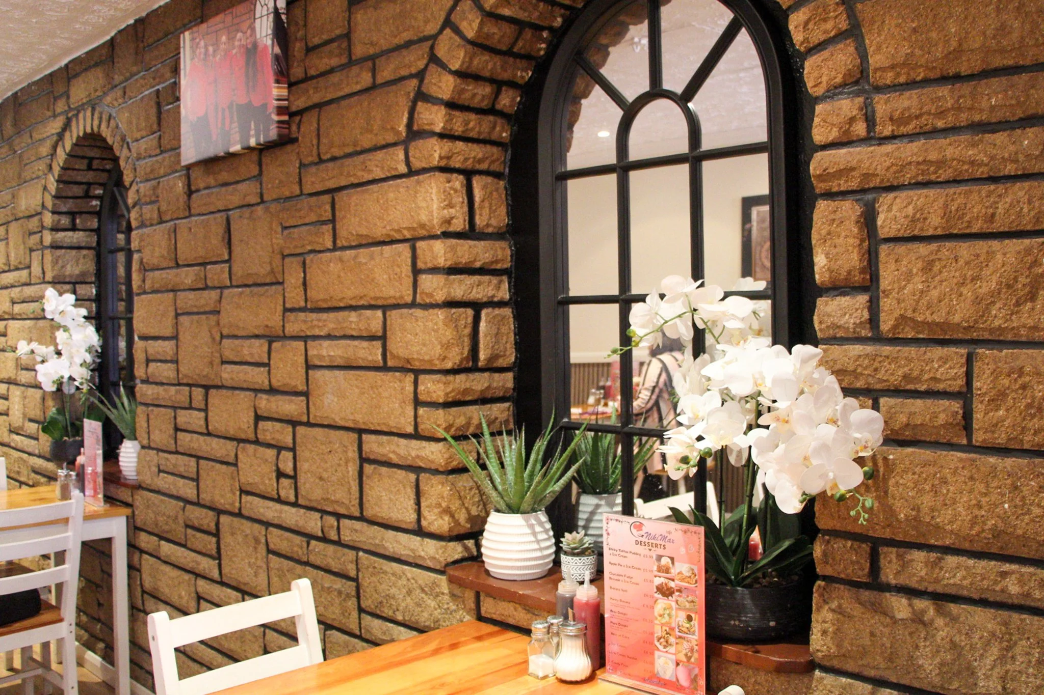 Café interior with wooden tables, white chairs, arched mirrors and plants against a stone wall.