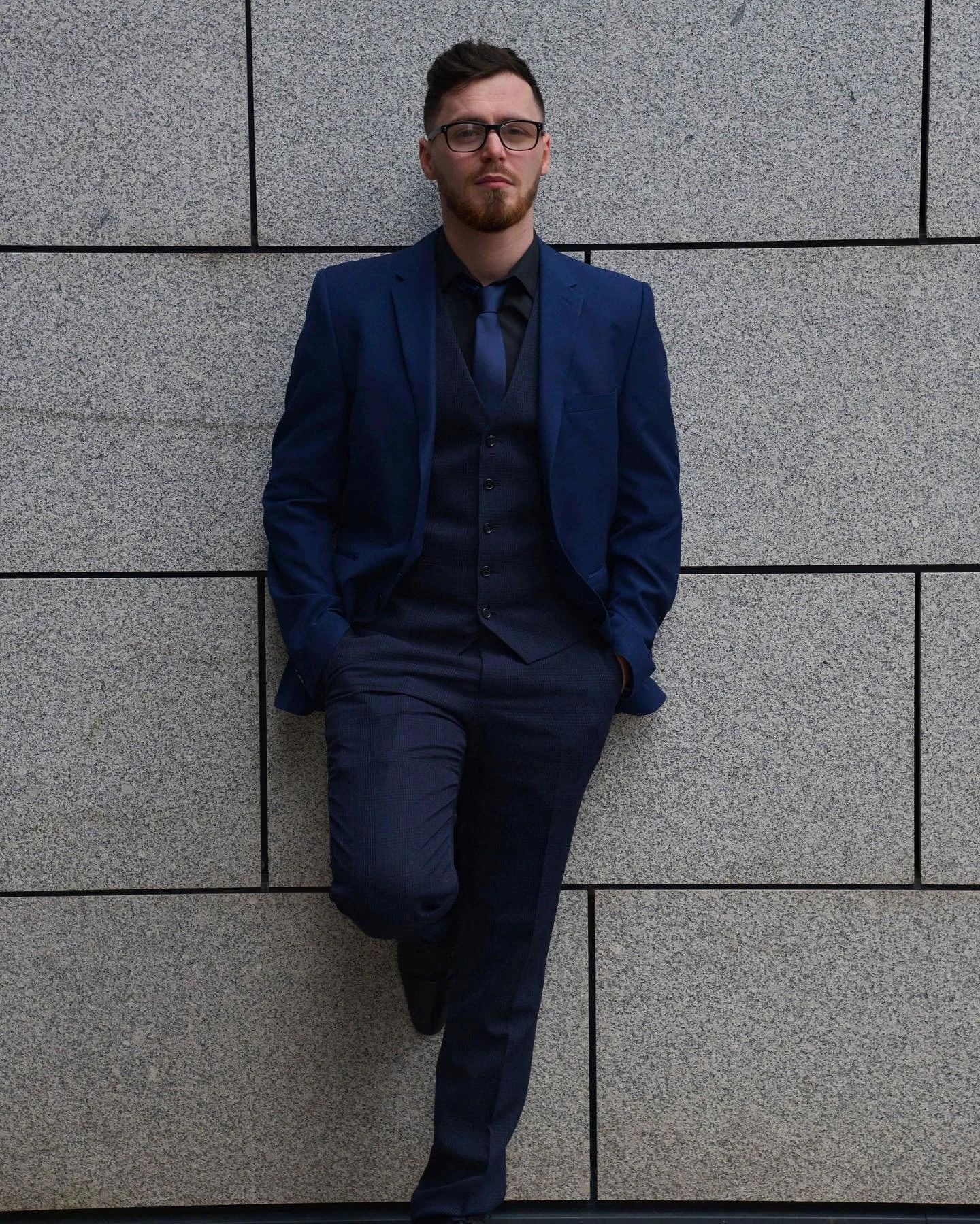 Man wearing glasses and a blue three-piece suit, posing against a grey stone wall.