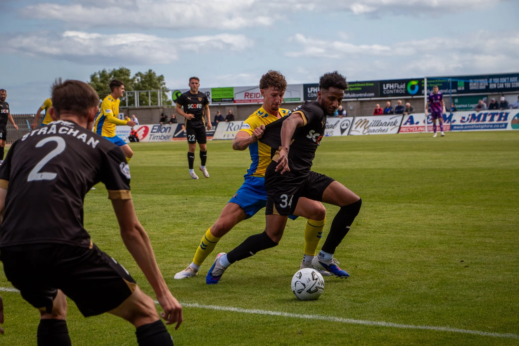Footballer in black shielding the ball from an opposing player in yellow and blue during a match.