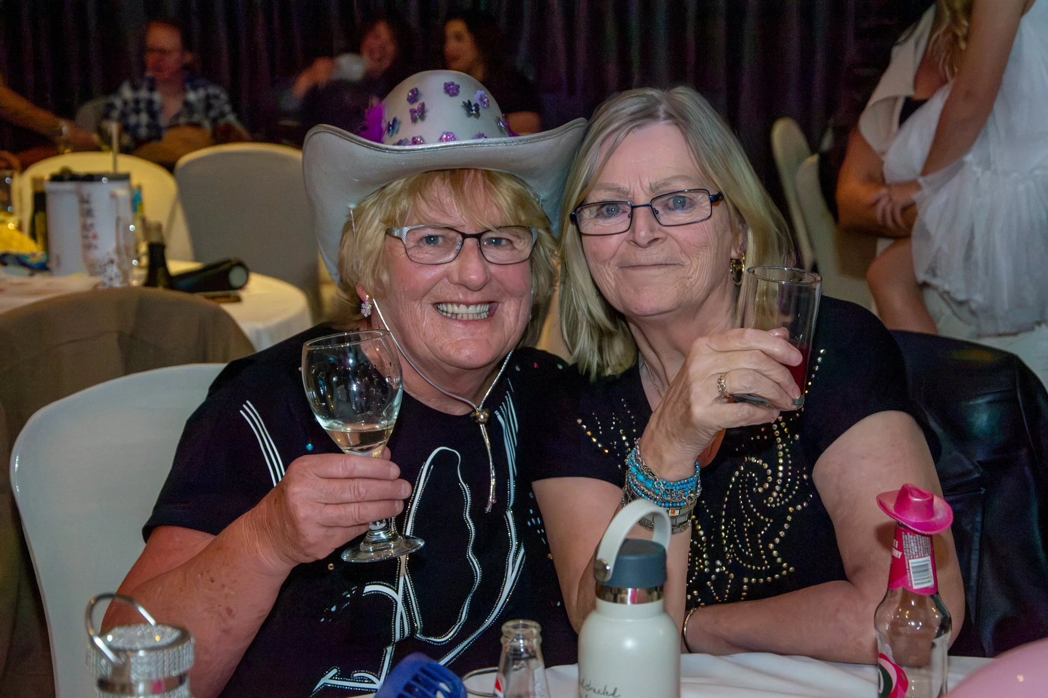 Two smiling guests holding drinks at a celebration, one wearing a decorated cowboy hat.