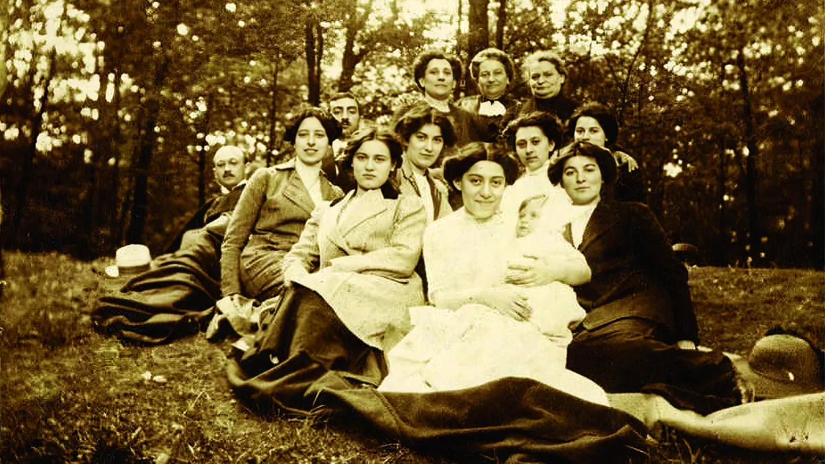 Group of women and a man outdoors in a forest, sitting and standing on the grass, some women holding a baby, with trees in the background.