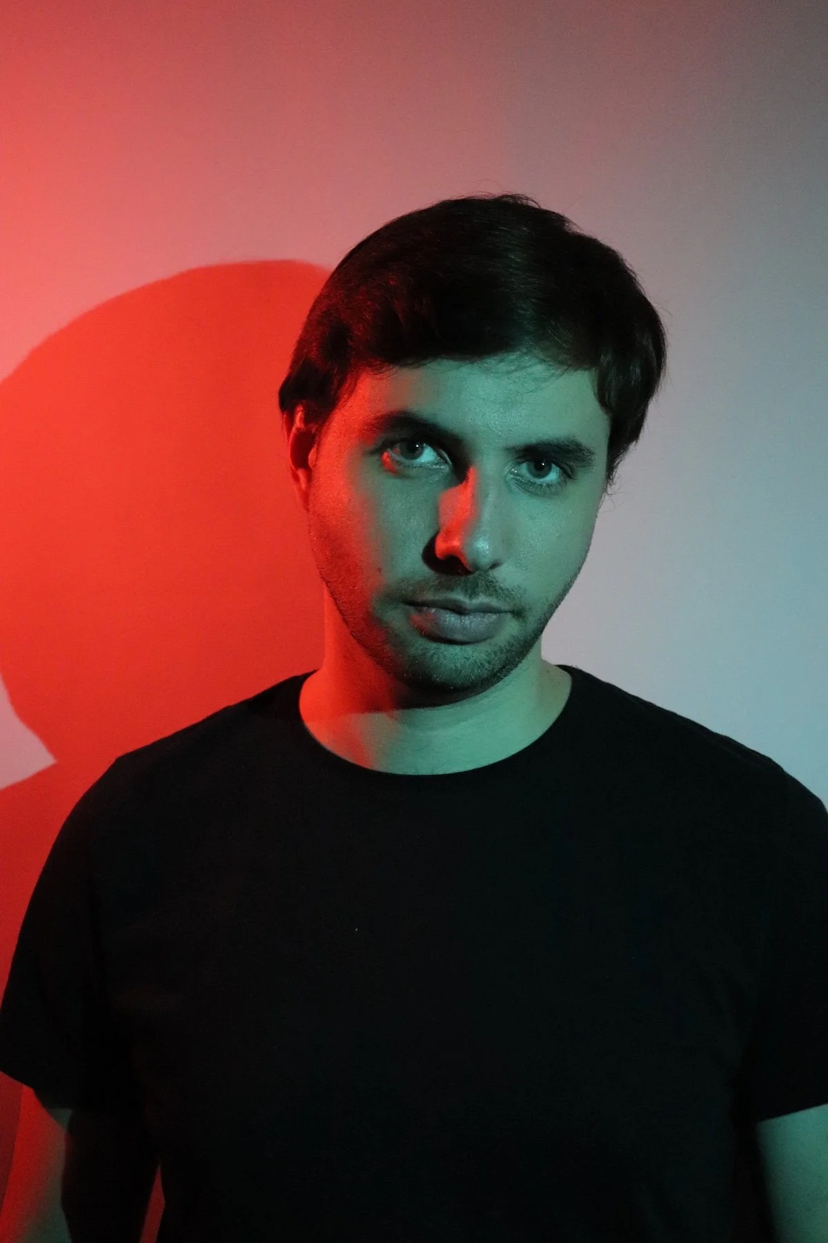 Jeune homme avec cheveu brun, regardant vers l'objectif, fond avec éclairage rouge et vert, portant un t-shirt noir.