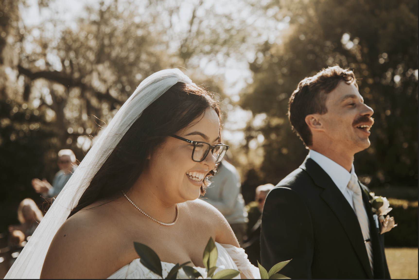 Happy bride and groom at outdoor wedding ceremony, the bride with glasses and veil, the groom in a suit and tie, with guests in the background.