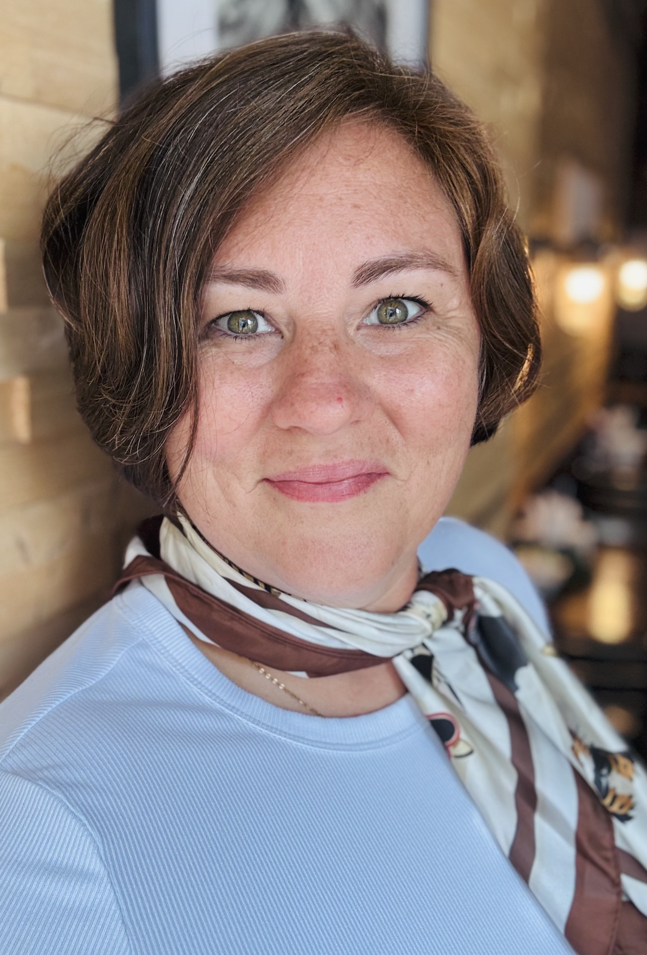 A woman with short brown hair and green eyes, wearing a light blue top and a patterned scarf, smiling softly, with a brick wall and warm lighting in the background.