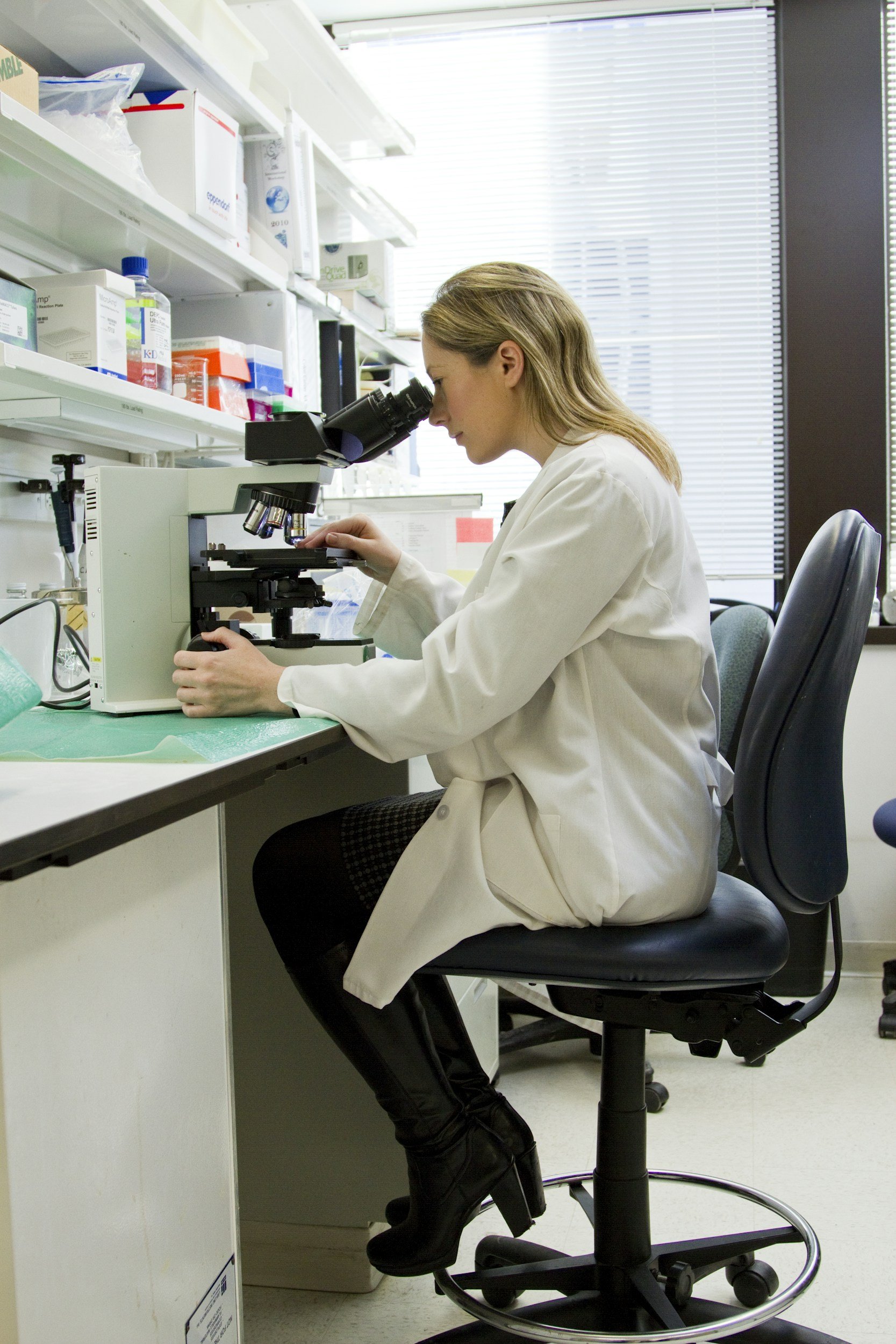 A female scientist looking into a microscope in a laboratory setting with shelves of lab supplies and equipment behind her.