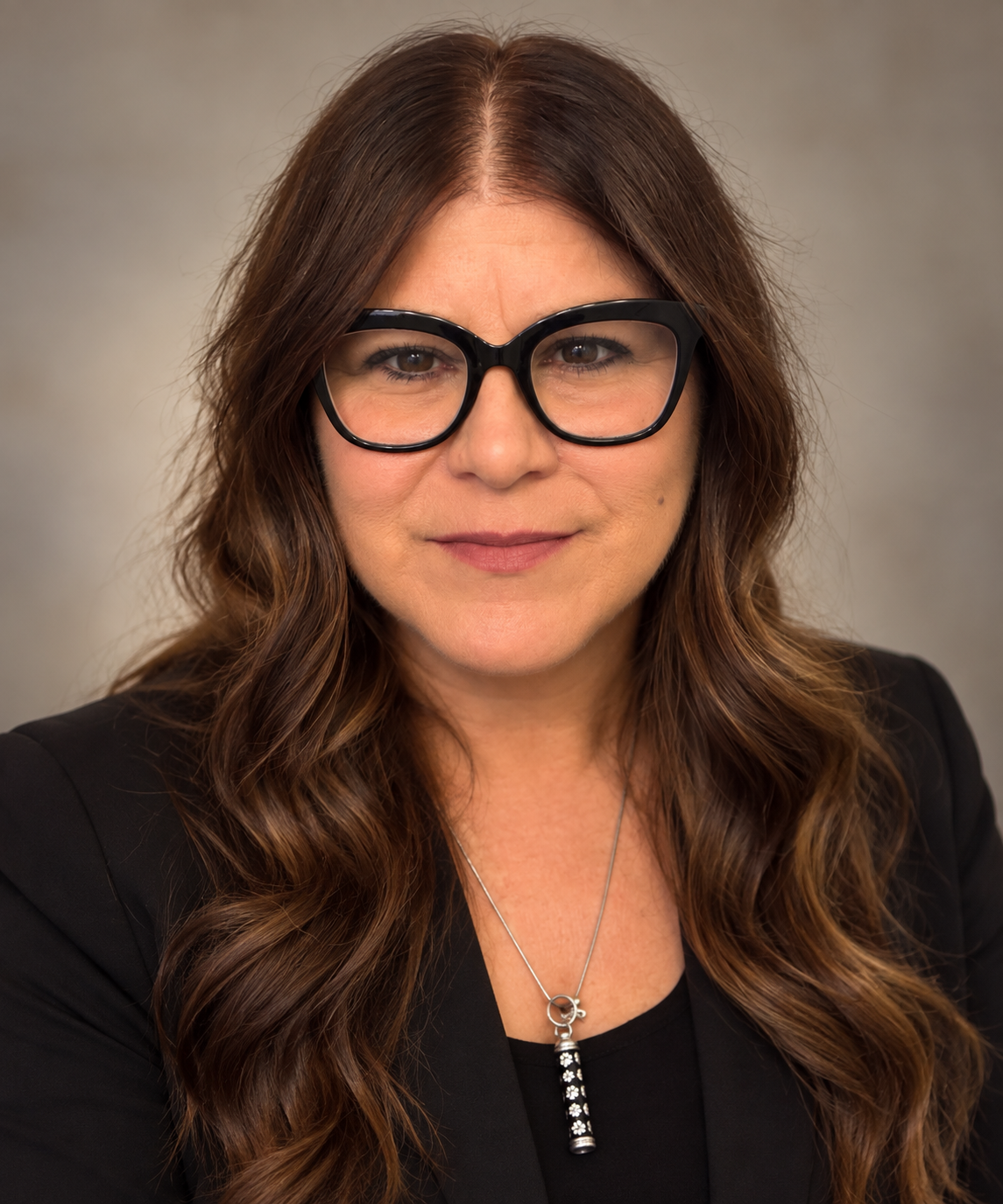 Close-up portrait of a woman with long wavy brown hair wearing black glasses, a black blazer, and a necklace with a black-and-white pendant, against a neutral background.